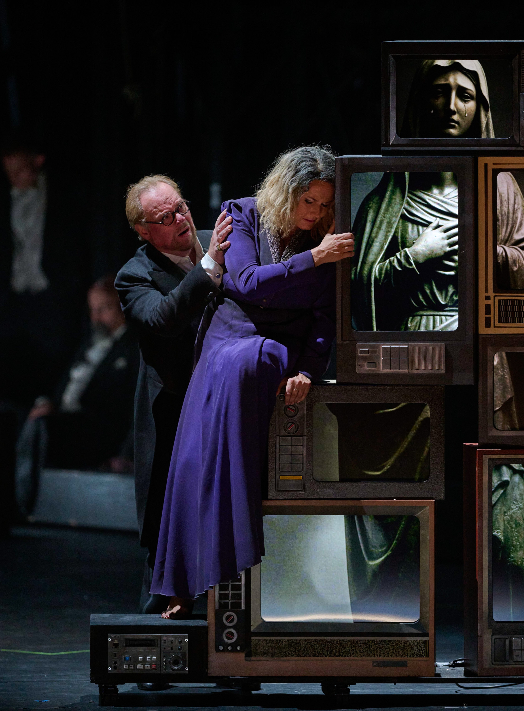 Eine Frau in einem violetten Kleid hält sich an einem Turm aus Fernsehern, die eine Statue zeigen, fest. Neben ihr kniet ein Mann.