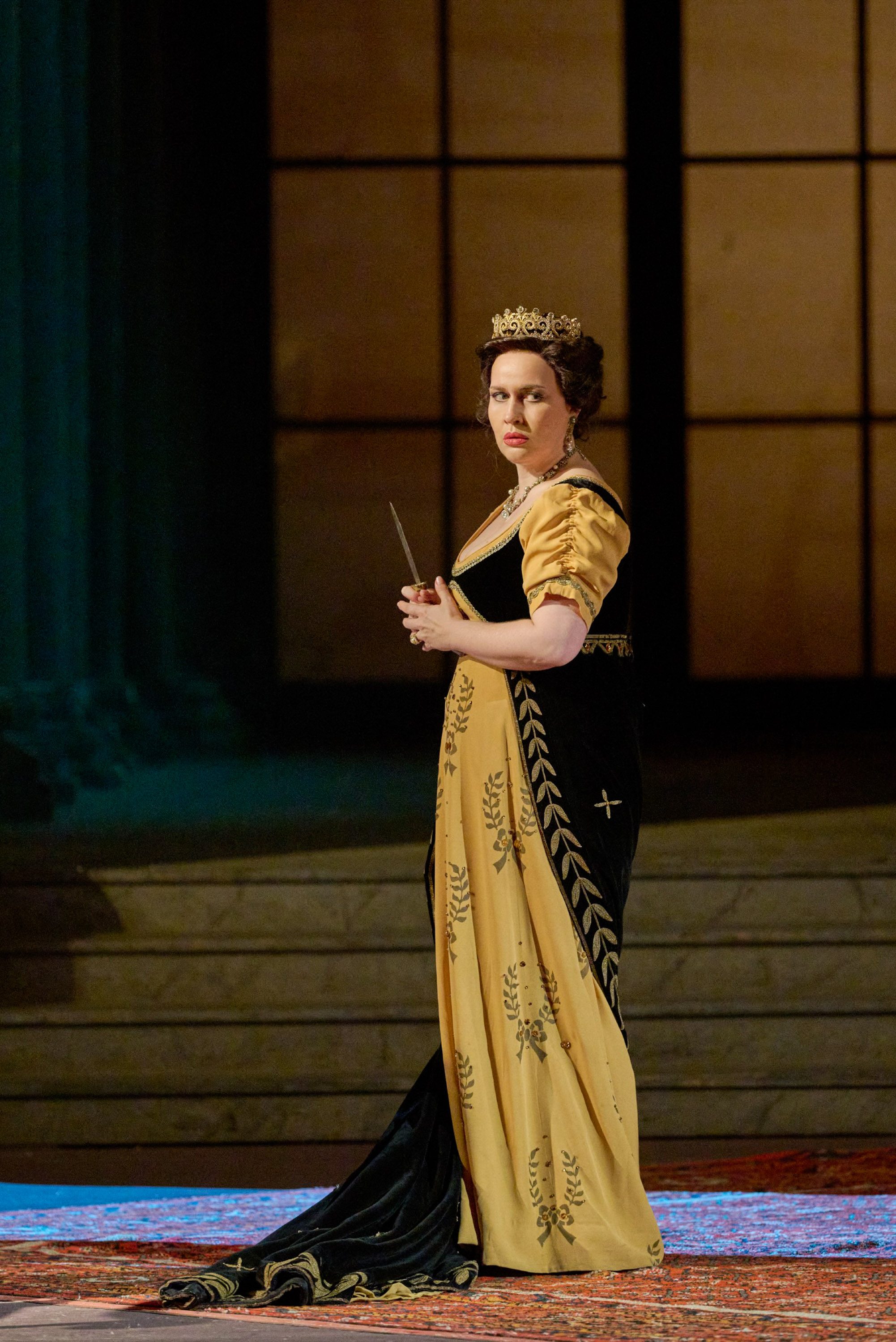 Eine Frau in einem schwarz-gelben Kleid und einer Krone auf dem Kopf hält einen Dolch in ihren Händen. 