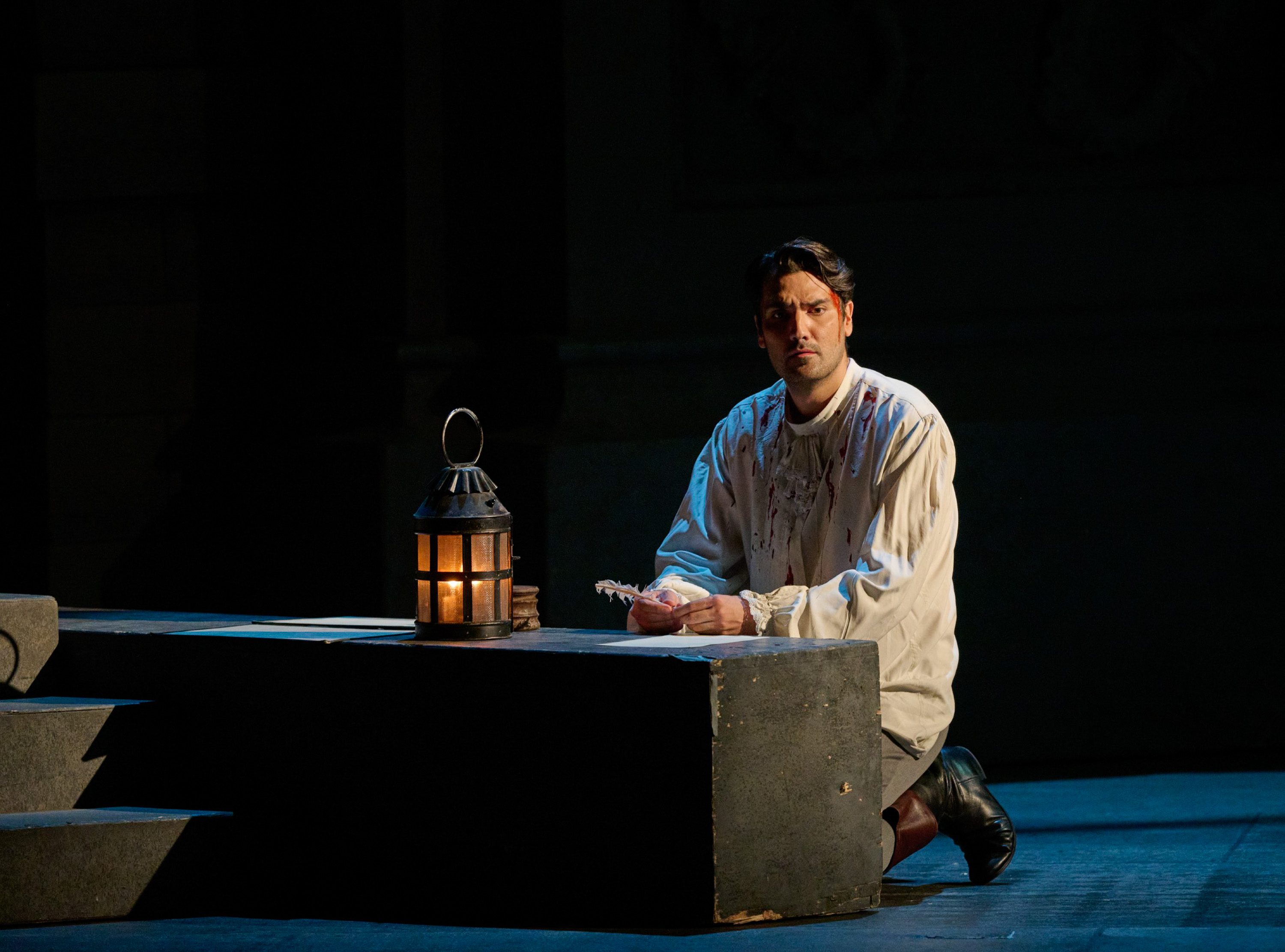 Ein Mann mit einer Platzwunde an der Schläfe kniet vor einem Sockel. Darauf steht eine Laterne, daneben liegt ein Blatt Papier. In der Hand hält der Mann eine Schreibfeder.