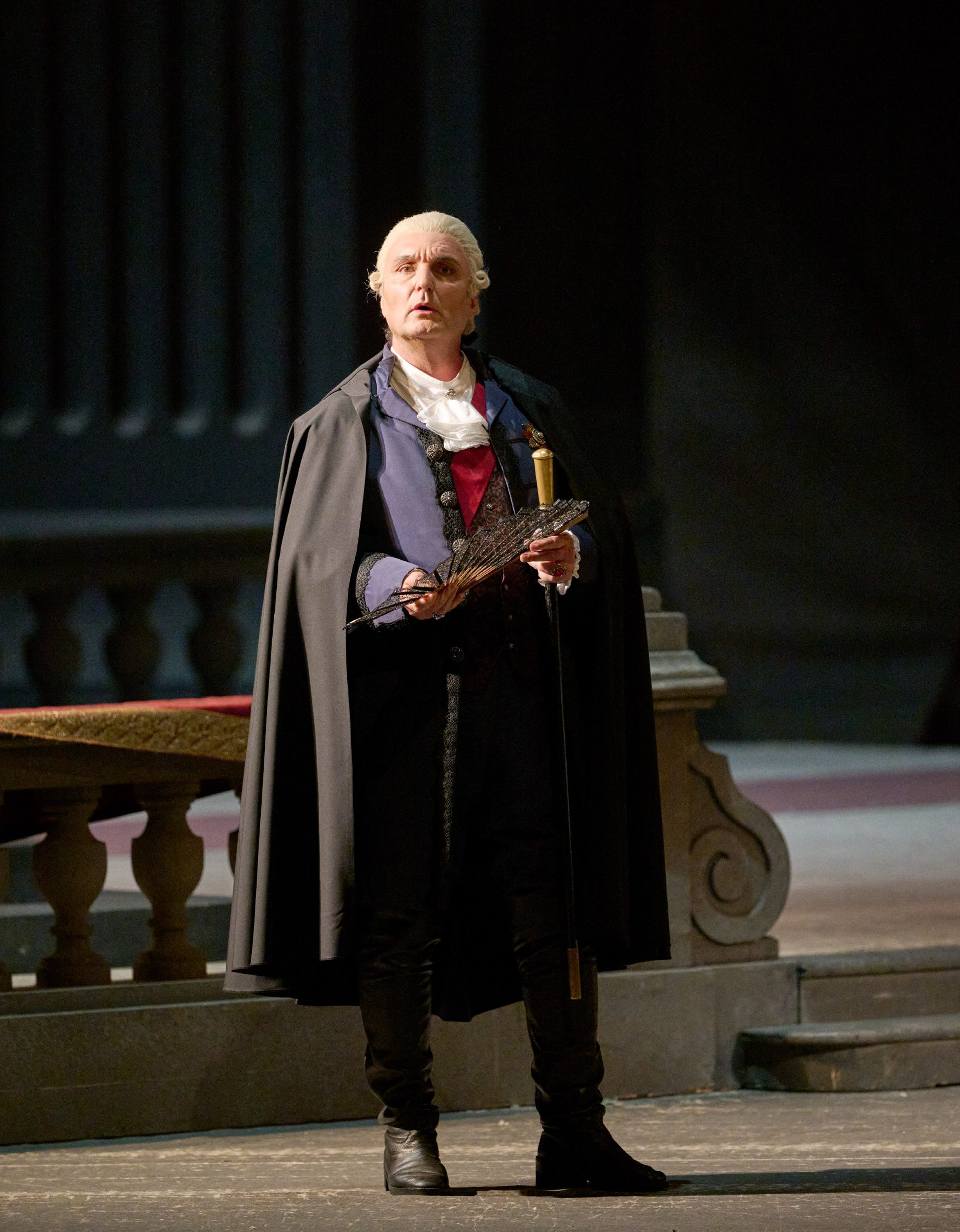 Man sieht einen Mann in einem dunklen Umhang, der vor einem dunklen Hintergrund steht. Er trägt eine weiße Perücke auf dem Kopf und hält einen Fächer in der Hand.