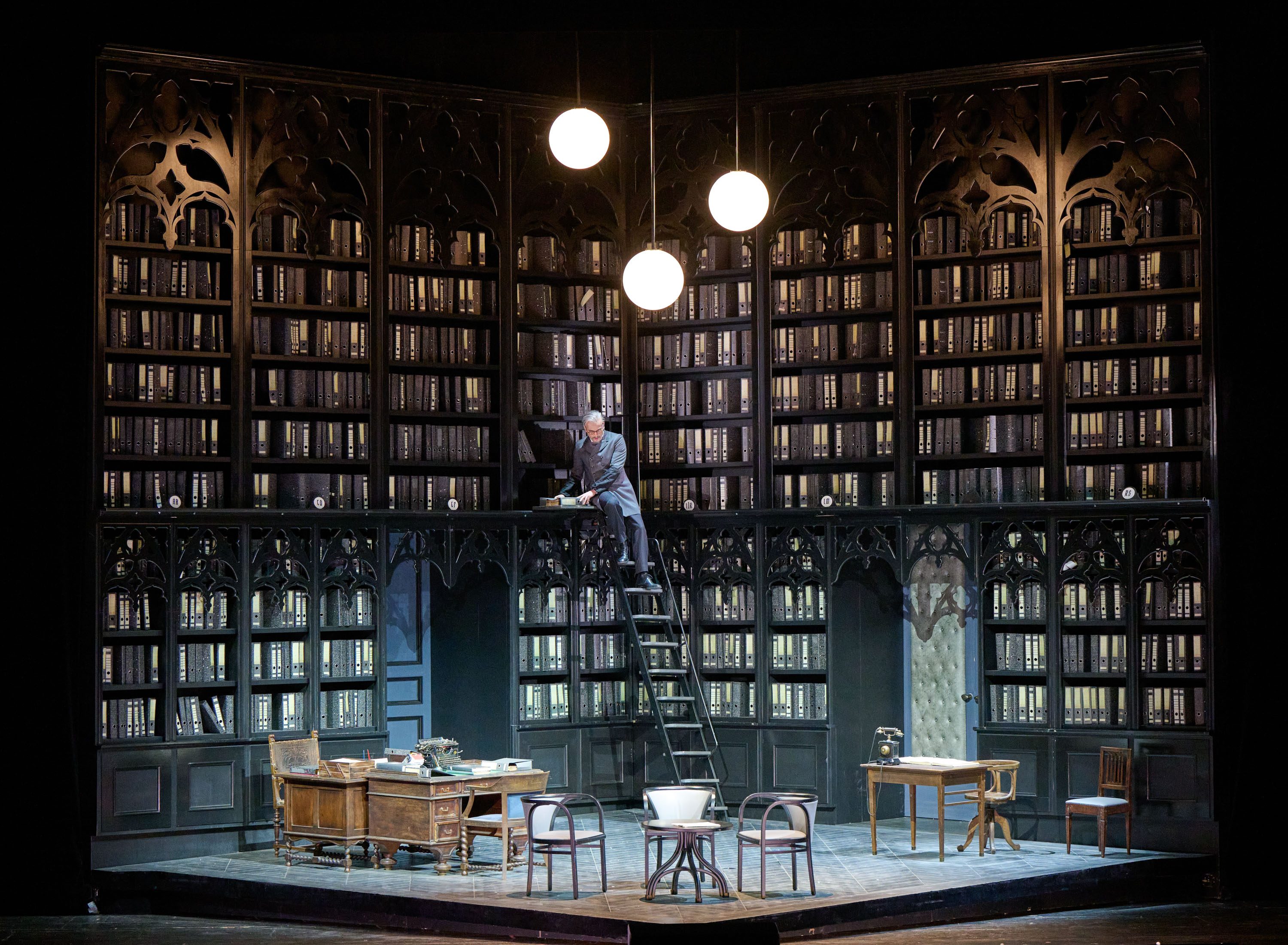 Eine Bühnenansicht mit einem hohen Bücherregal, das voll mit Aktenordnern gefüllt ist. Ein Mann sitzt auf einer Leiter und schaut auf einen Tisch mit Schreibmaschine und Büchern. Auf der Bühne sind außerdem mehrere Stühle und ein weiterer Tisch zu sehen.