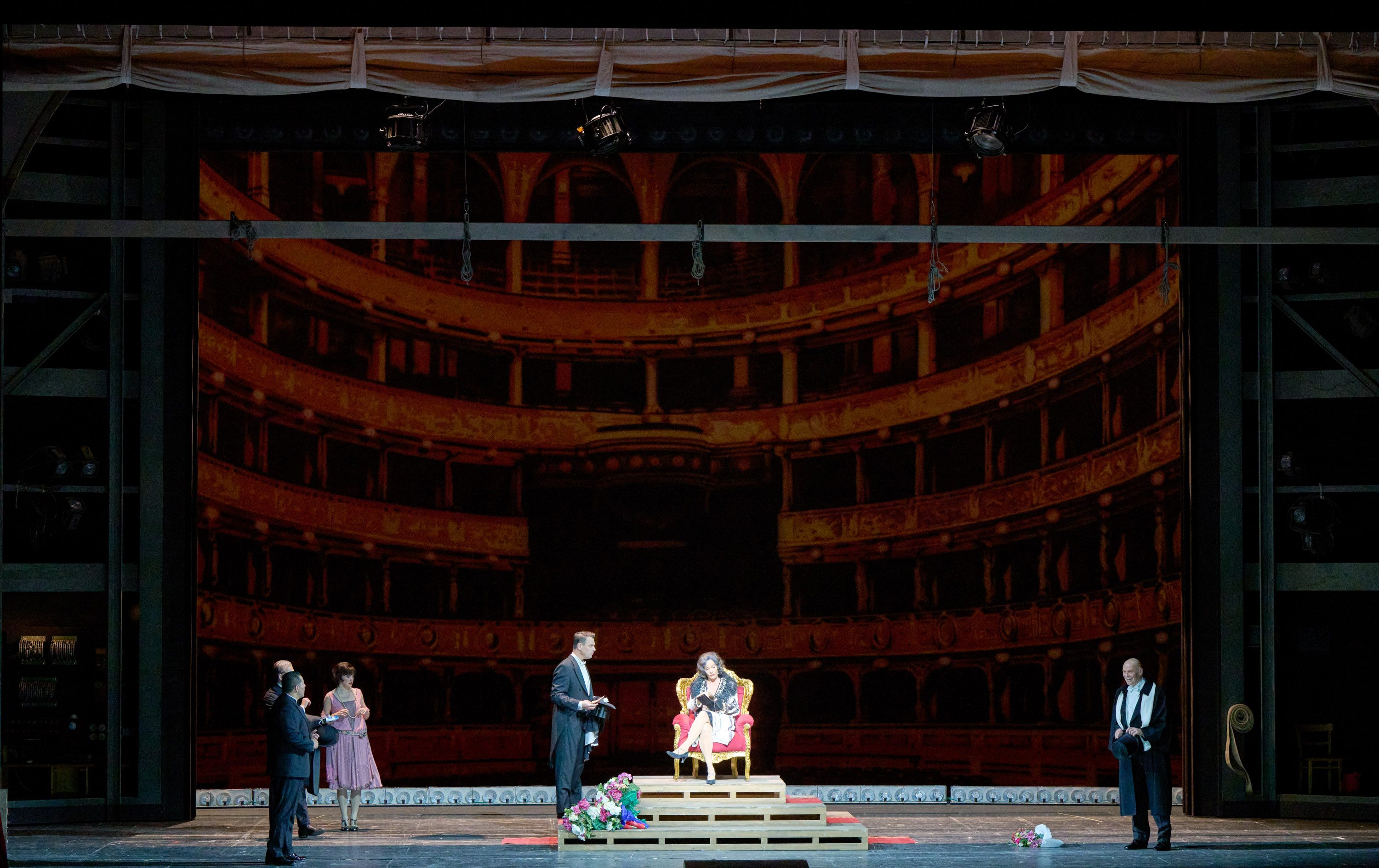 Auf der Bühne eines Theaters stehen mehrere Personen. Eine Frau sitzt auf einem Thron, umgeben von Blumen. Ein Mann im Anzug liest etwas vor, während andere Zuschauer aufmerksam zuschauen. Im Hintergrund ist eine imposante, verschnörkelte Theaterarchitektur sichtbar.