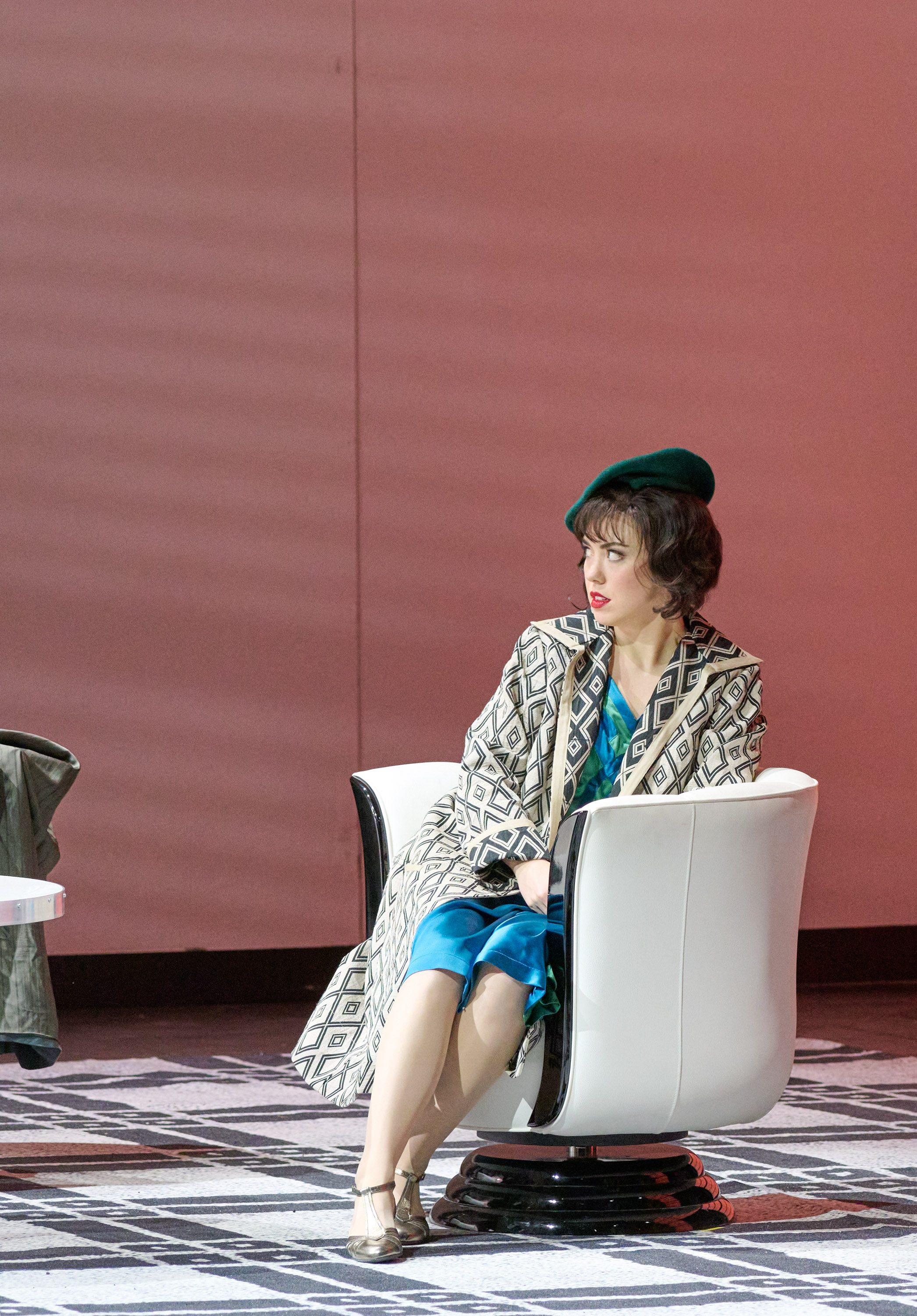 Eine Frau mit einem grünen Hut sitzt in einem modernen, weißen Stuhl. Sie trägt einen auffälligen Mantel mit geometrischem Muster und ein blaues Kleid. Der Hintergrund ist in sanften Rottönen gehalten, während der Boden mit einem gemusterten Teppich bedeckt ist.