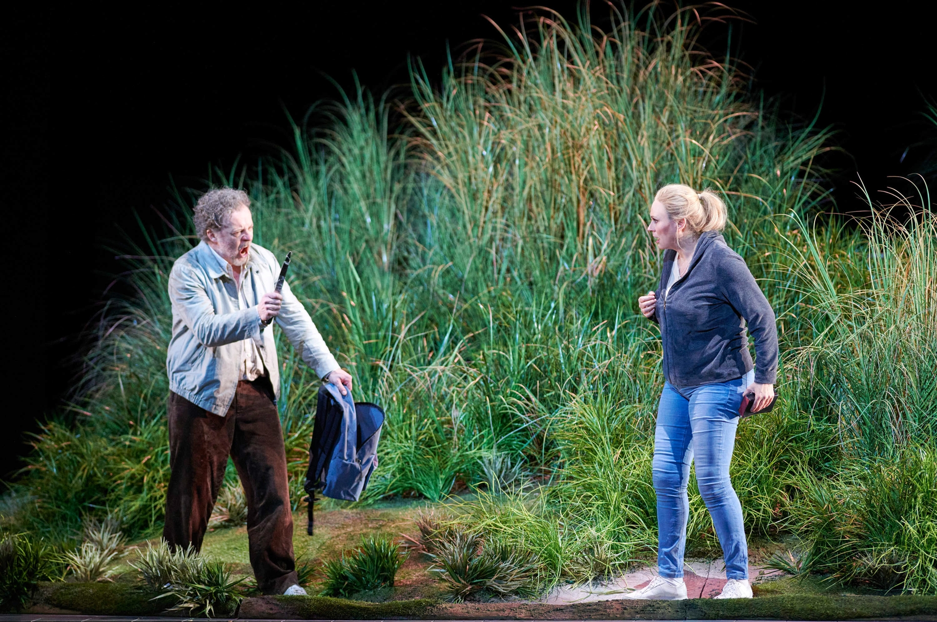 Ein Mann und eine Frau stehen in einer Graslandschaft; er hält einen Rucksack in der einen und ein Messer in der anderen Hand.