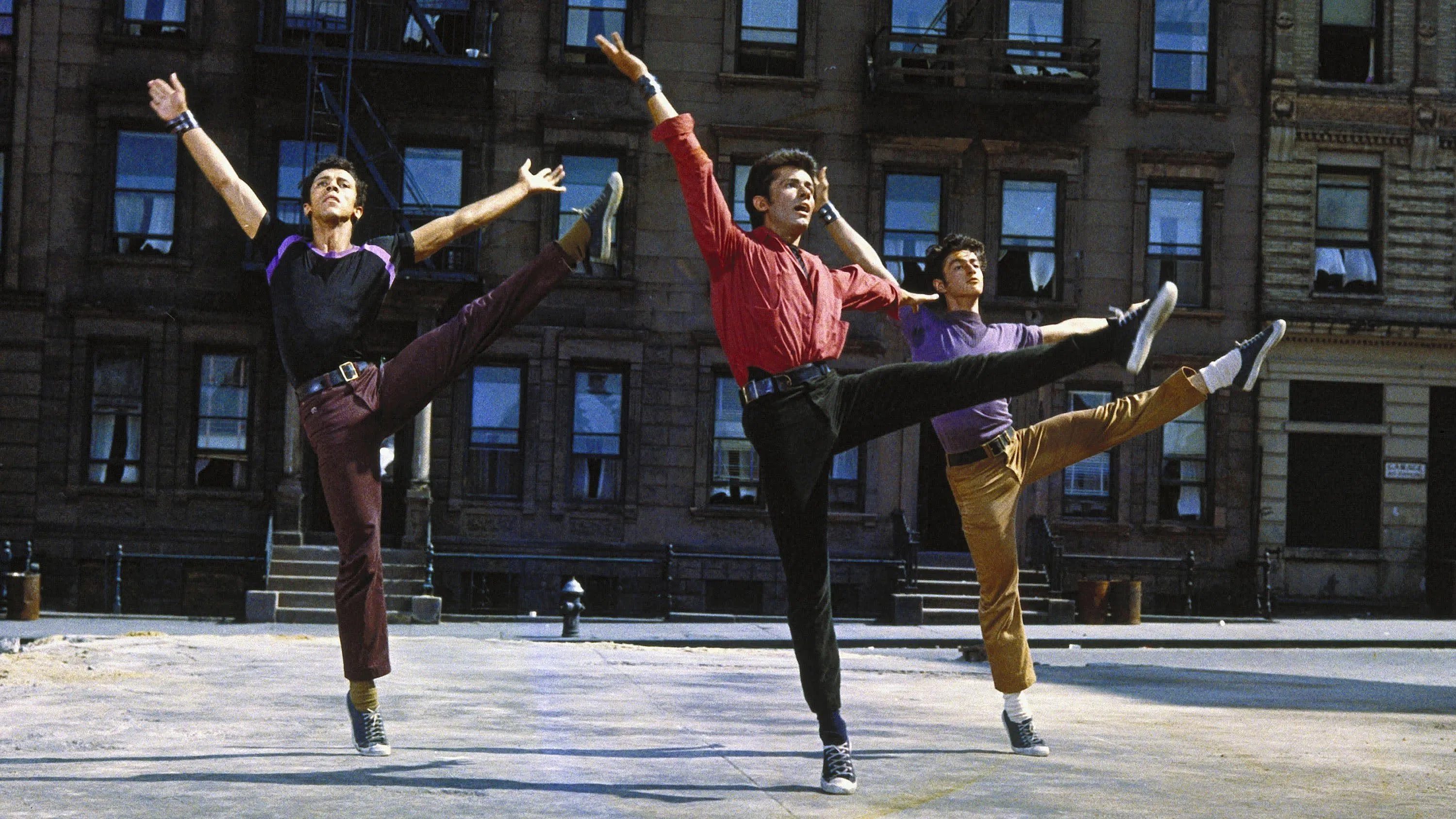 Three male dancers are performing a synchronized dance routine outdoors, displaying dynamic leg kicks. They are dressed in colorful shirts and pants, showcasing enthusiasm and energy. The backdrop features a city building with numerous windows.