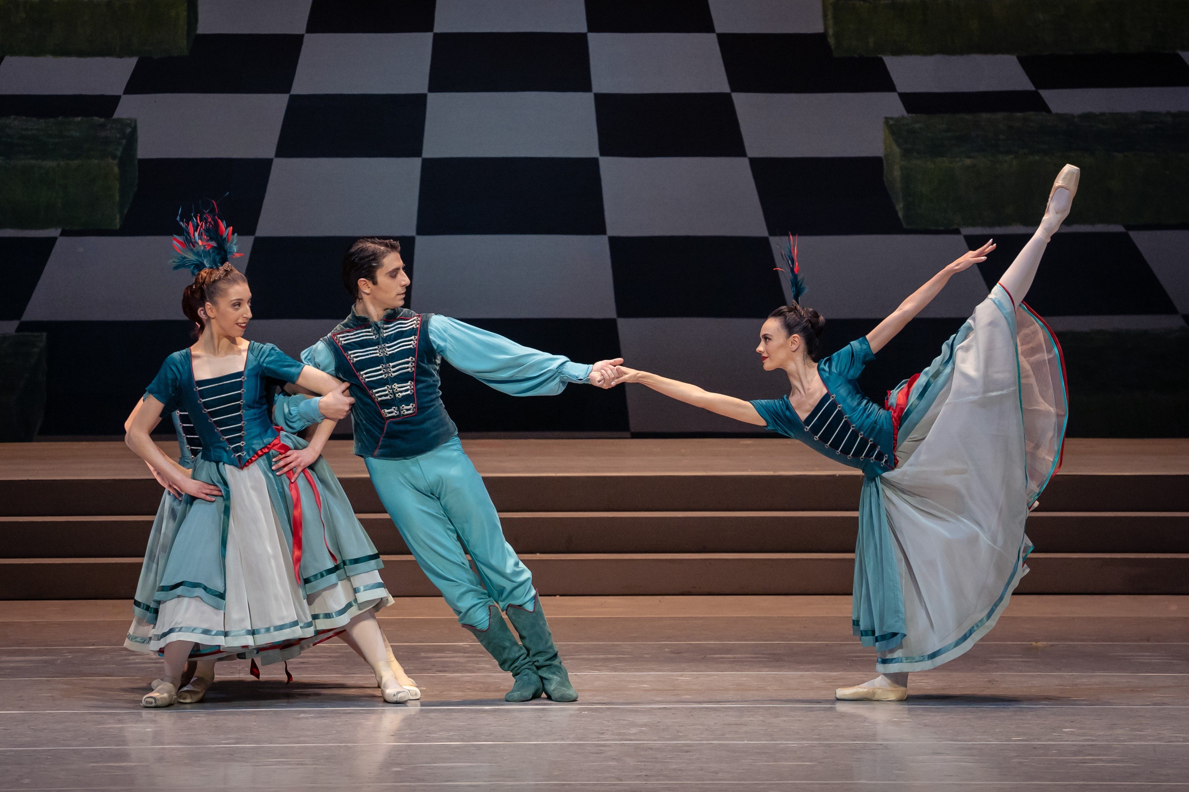 Drei Tänzer in eleganter Ballettkleidung führen eine Choreografie auf einer Bühne aus. Sie zeigen unterschiedliche Posen, wobei einer einen Partner hält, während die andere einen hohen arabesque vollführt. Der Hintergrund ist in Schwarz-Weiß-Klötzen gestaltet.