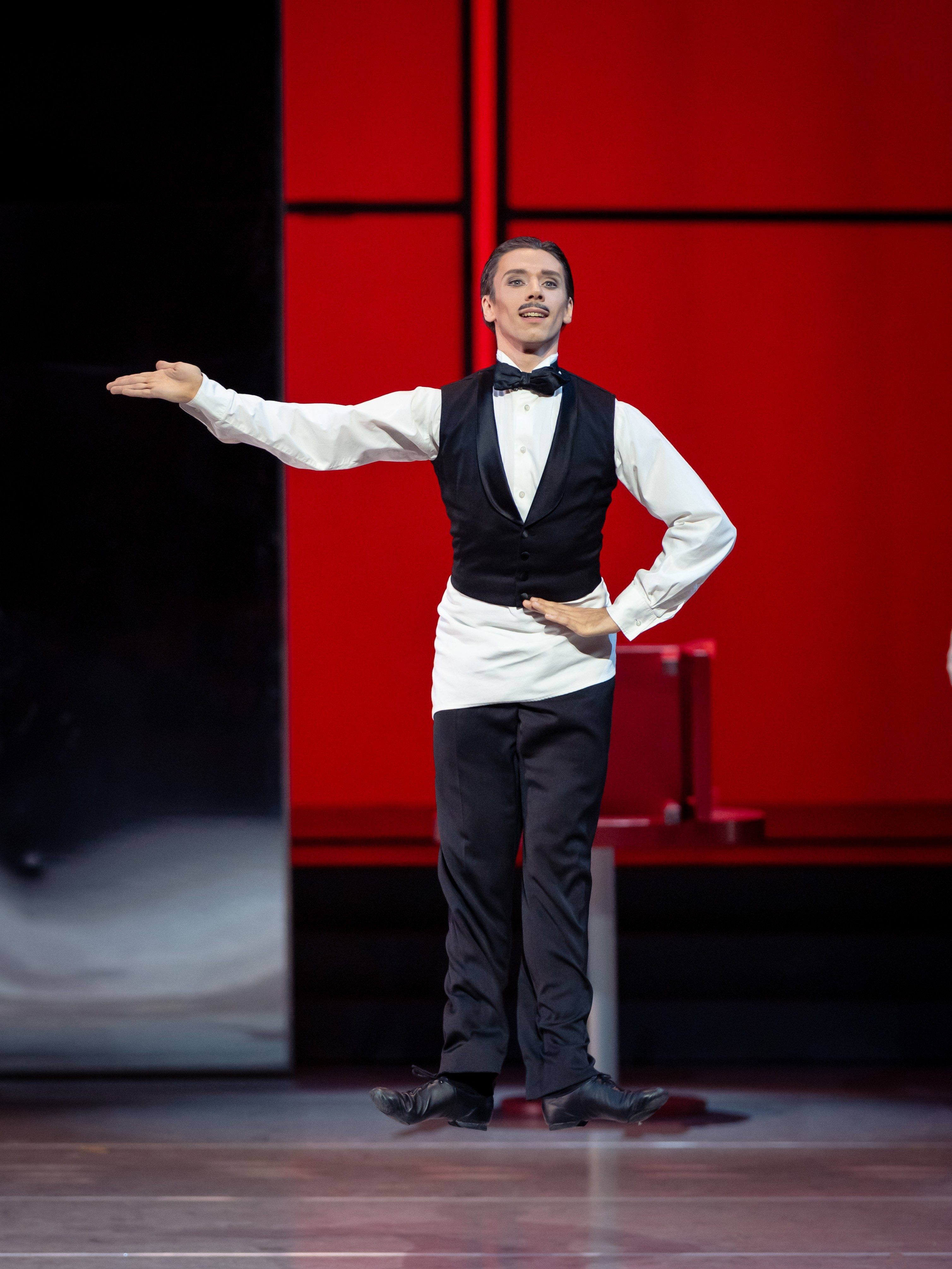 A dancer in a black vest and white dress shirt performs on stage, striking a graceful pose with one arm extended. The background features a vibrant red color, enhancing the dramatic performance. The dancer exudes elegance and confidence.