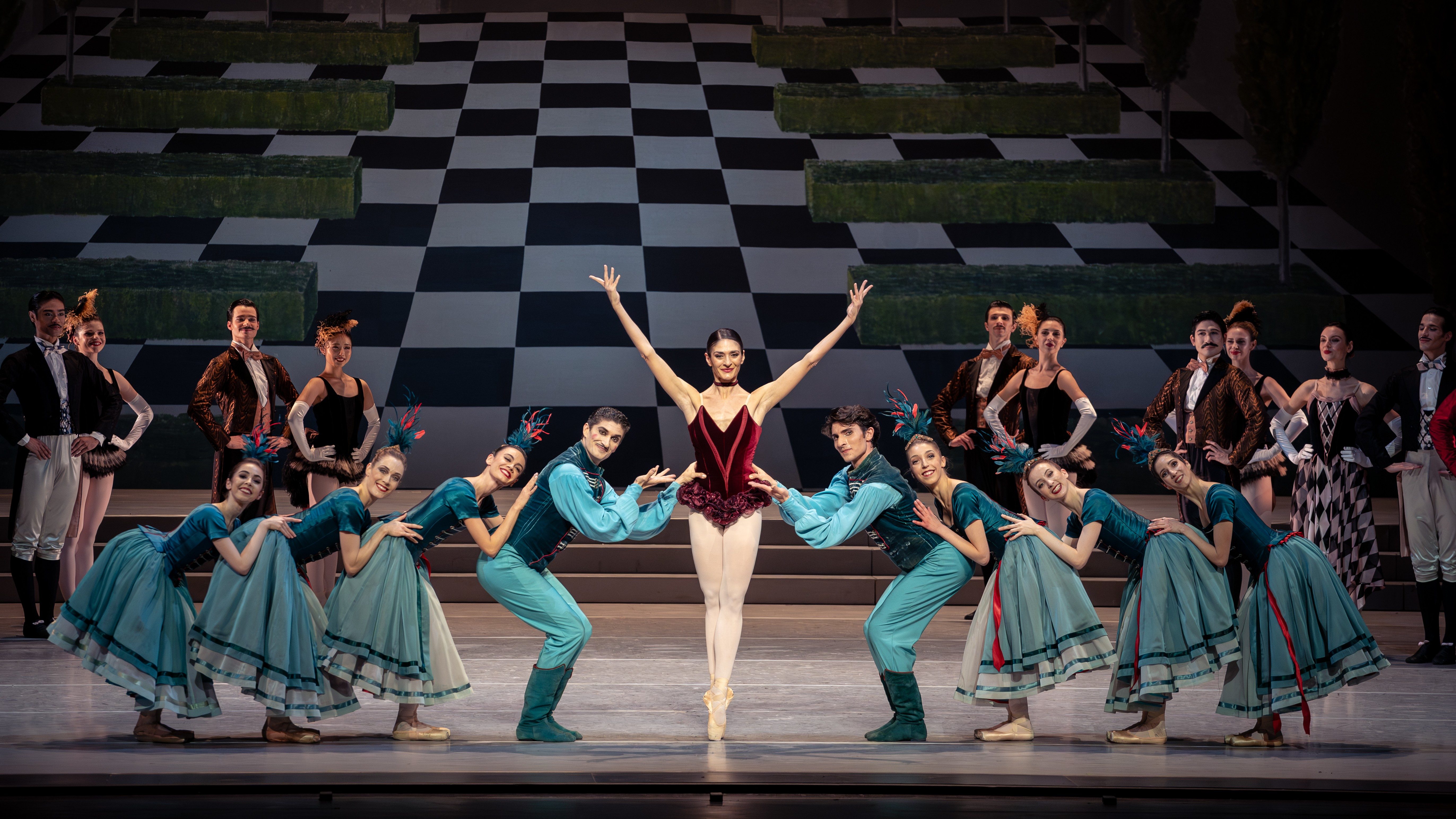 Eine Ballettaufführung zeigt eine Tänzerin in einem roten Kostüm, die in der Mitte des Bühnenraums posiert. Um sie herum stehen mehrere Tänzer in blauen Kostümen, die sie umschmeicheln und verschiedene Posen einnehmen. Der Hintergrund ist mit geometrischen Mustern dekoriert.