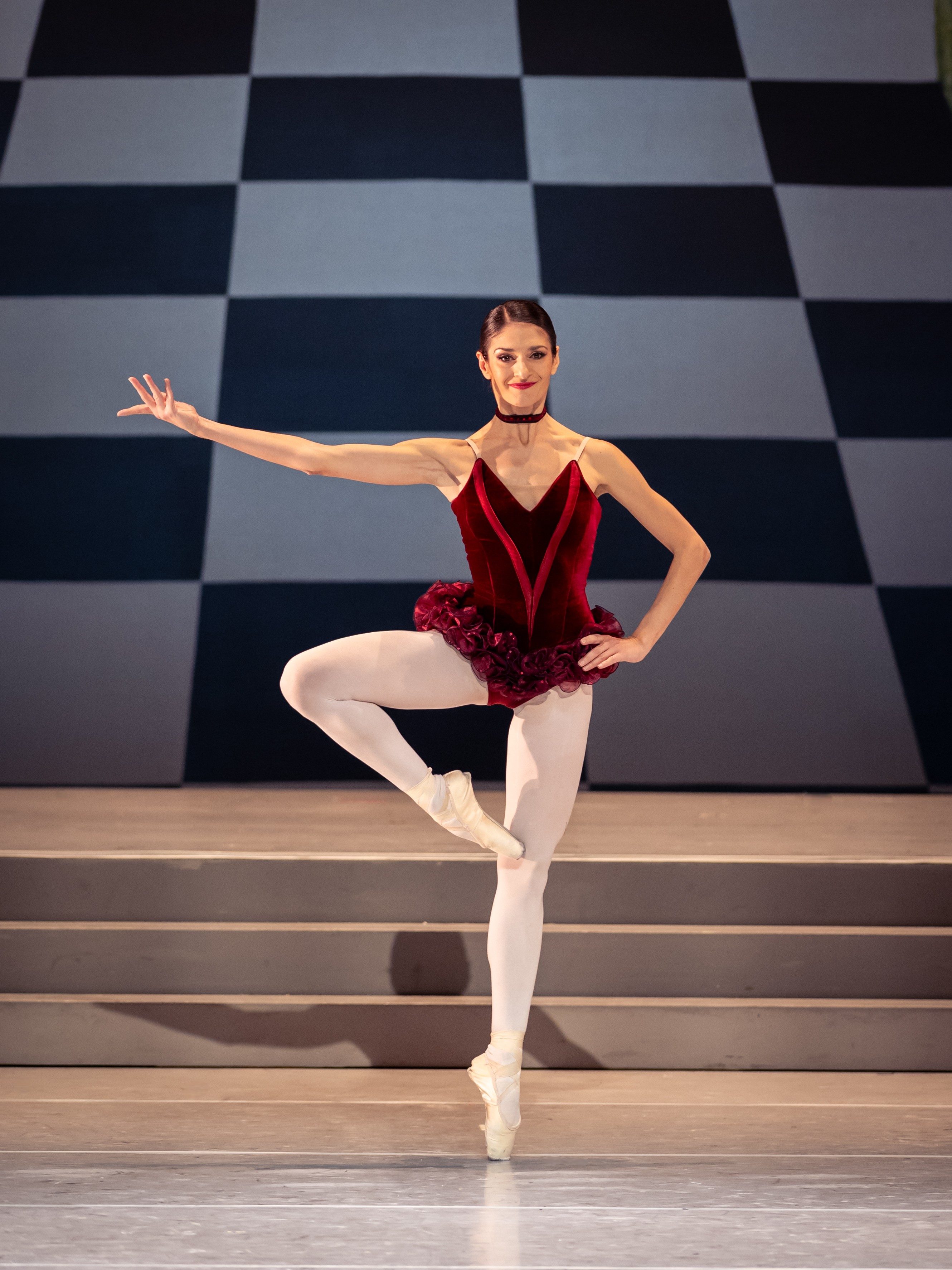 Eine Balletttänzerin in einem roten Kostüm präsentiert eine Pose auf einem Bein, während sie einen Arm elegant zur Seite ausstreckt. Der Hintergrund zeigt ein Schachbrettmuster, das die dynamische Atmosphäre der Darbietung unterstreicht.