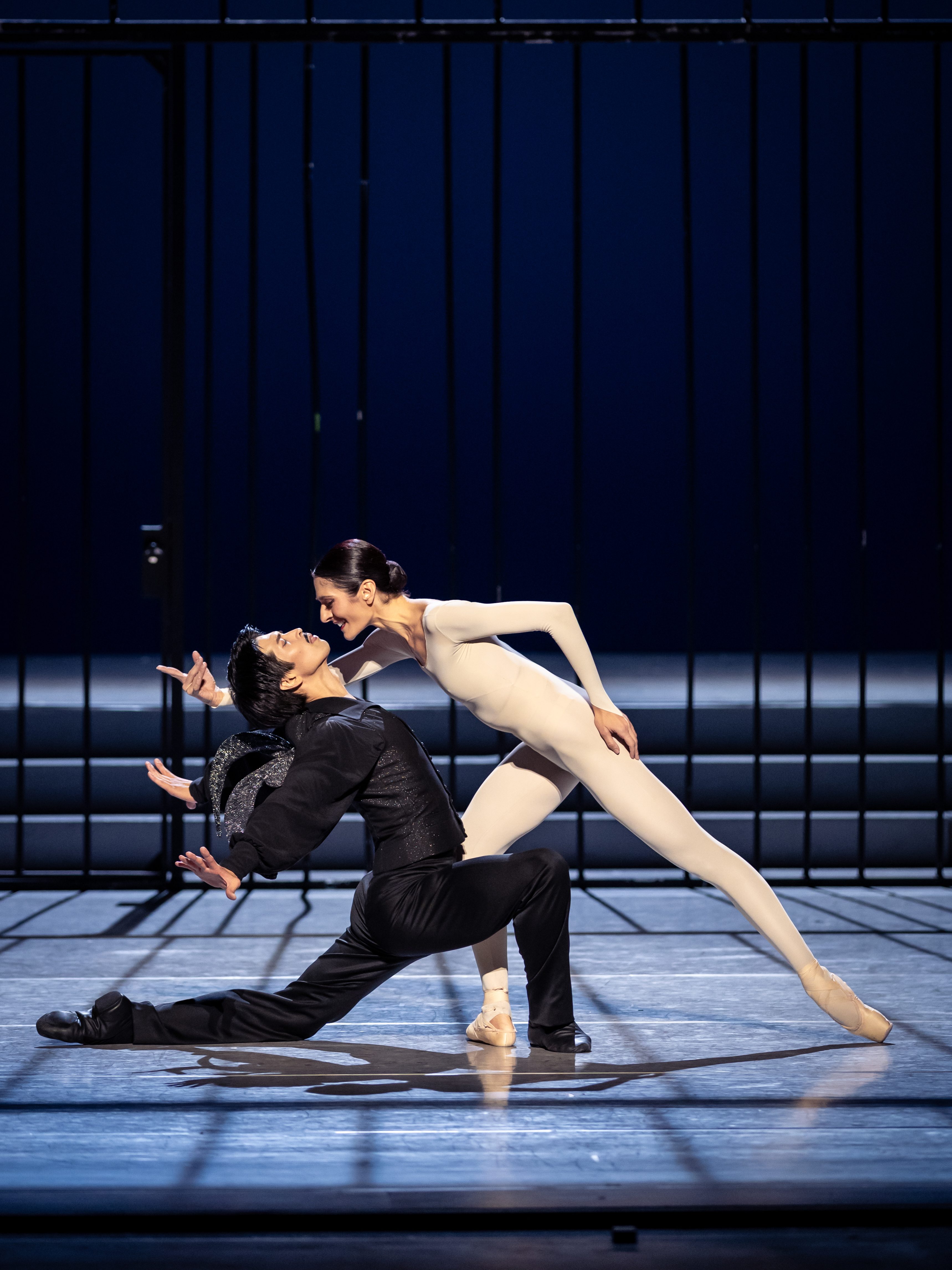 Ein männlicher Tänzer in schwarzer Kleidung und ein weiblicher Tänzer in einem hautengen weißen Anzug führen einen ausdrucksstarken Tanz auf. Sie sind nah beieinander, die Körperhaltung zeigt Leidenschaft und Dynamik. Der Hintergrund ist dunkel und verstärkt die Intensität ihrer Darbietung.