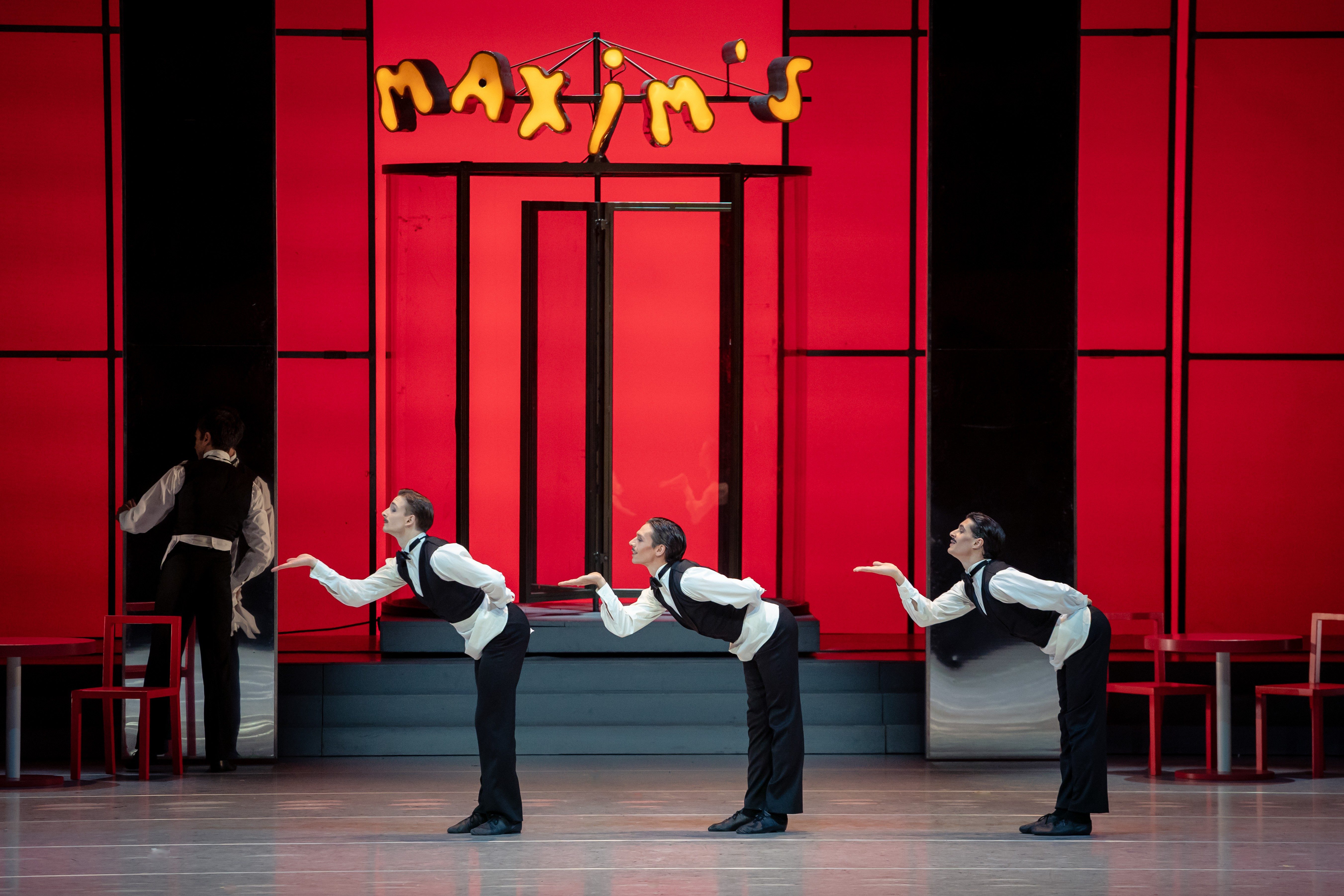 Drei Tänzer in eleganter Kleidung stehen in einer geschwungenen Formation. Sie neigen sich nach vorne und machen einladende Gesten. Im Hintergrund steht ein roter Wand mit dem Schriftzug „Maxim’s“. Die Szene vermittelt eine lebhafte, fröhliche Atmosphäre.