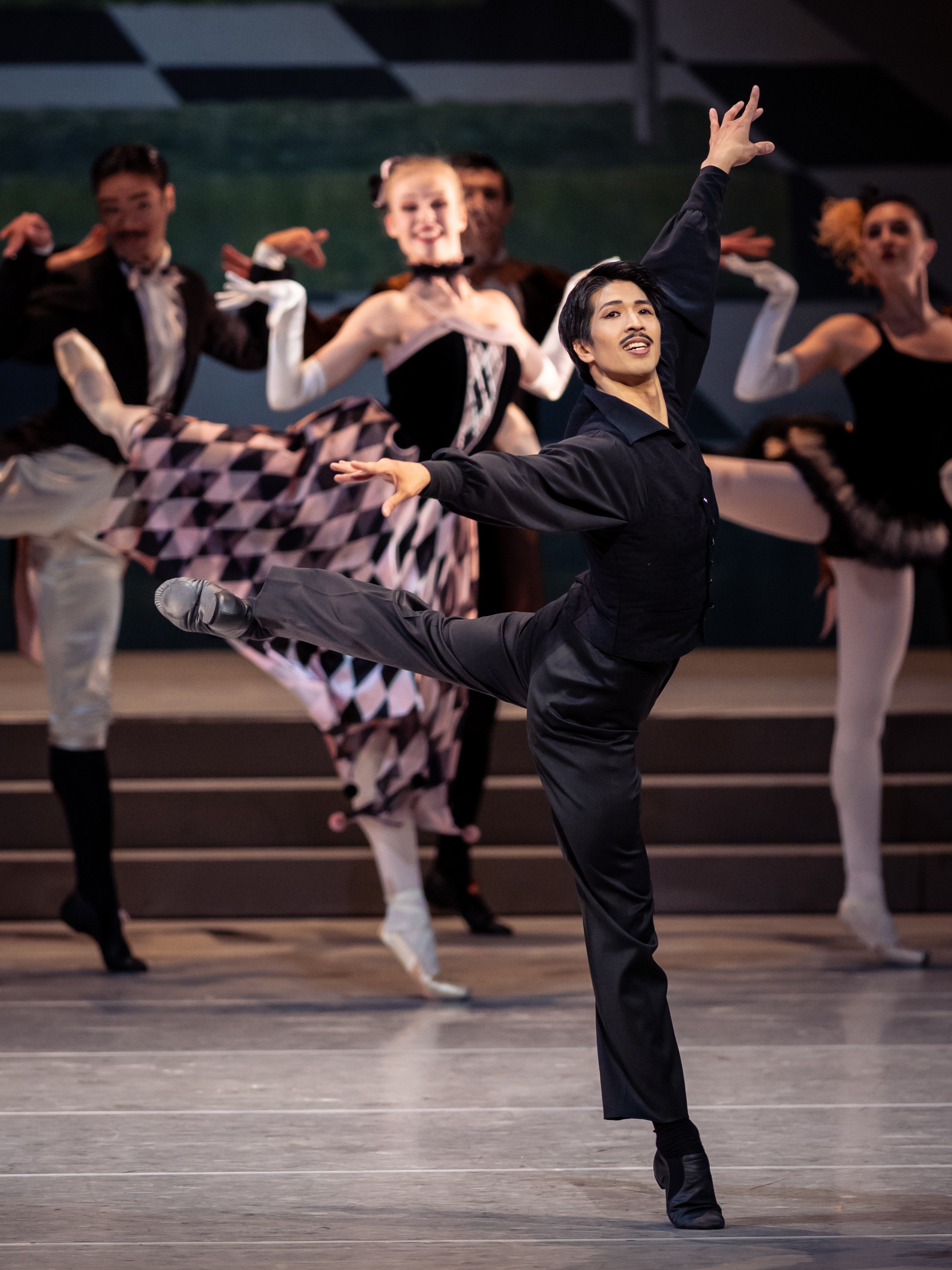 Eine Gruppe von Balletttänzern in vielfältigen Kostümen, die auf einer Bühne tanzen. Der vorderste Tänzer hebt elegant ein Bein und streckt einen Arm nach oben, während andere Tänzer verschiedene Posen einnehmen. Die Szene vermittelt Dynamik und Ausdruckskraft.