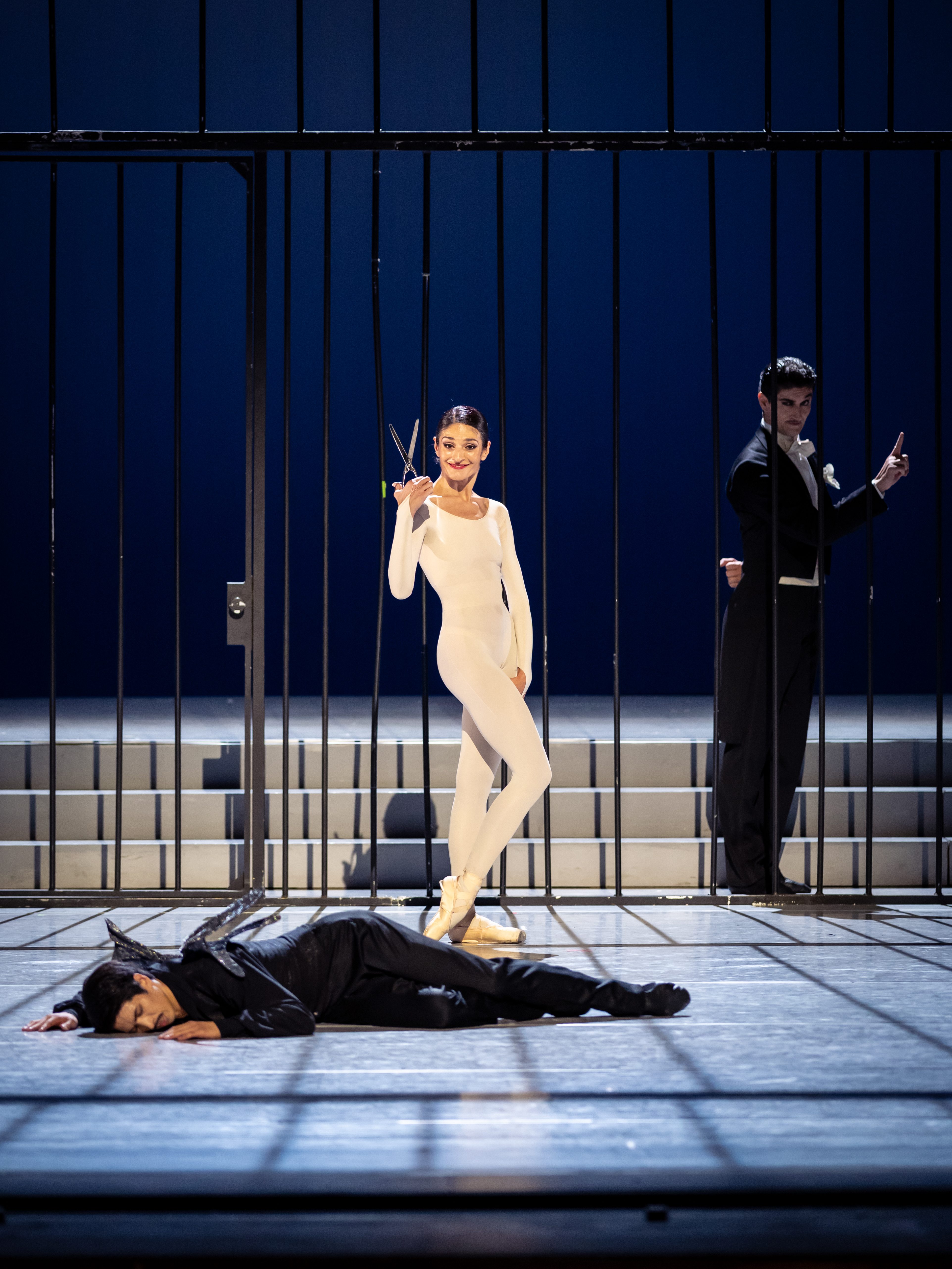 Eine Tänzerin in einem weißen Kostüm hält eine große Schere und lächelt, während sie zwischen zwei Gittern steht. Im Hintergrund liegt ein weiterer Tänzer in schwarzer Kleidung regungslos am Boden. Ein weiterer Mann im Smoking steht rechts und beobachtet die Szene.