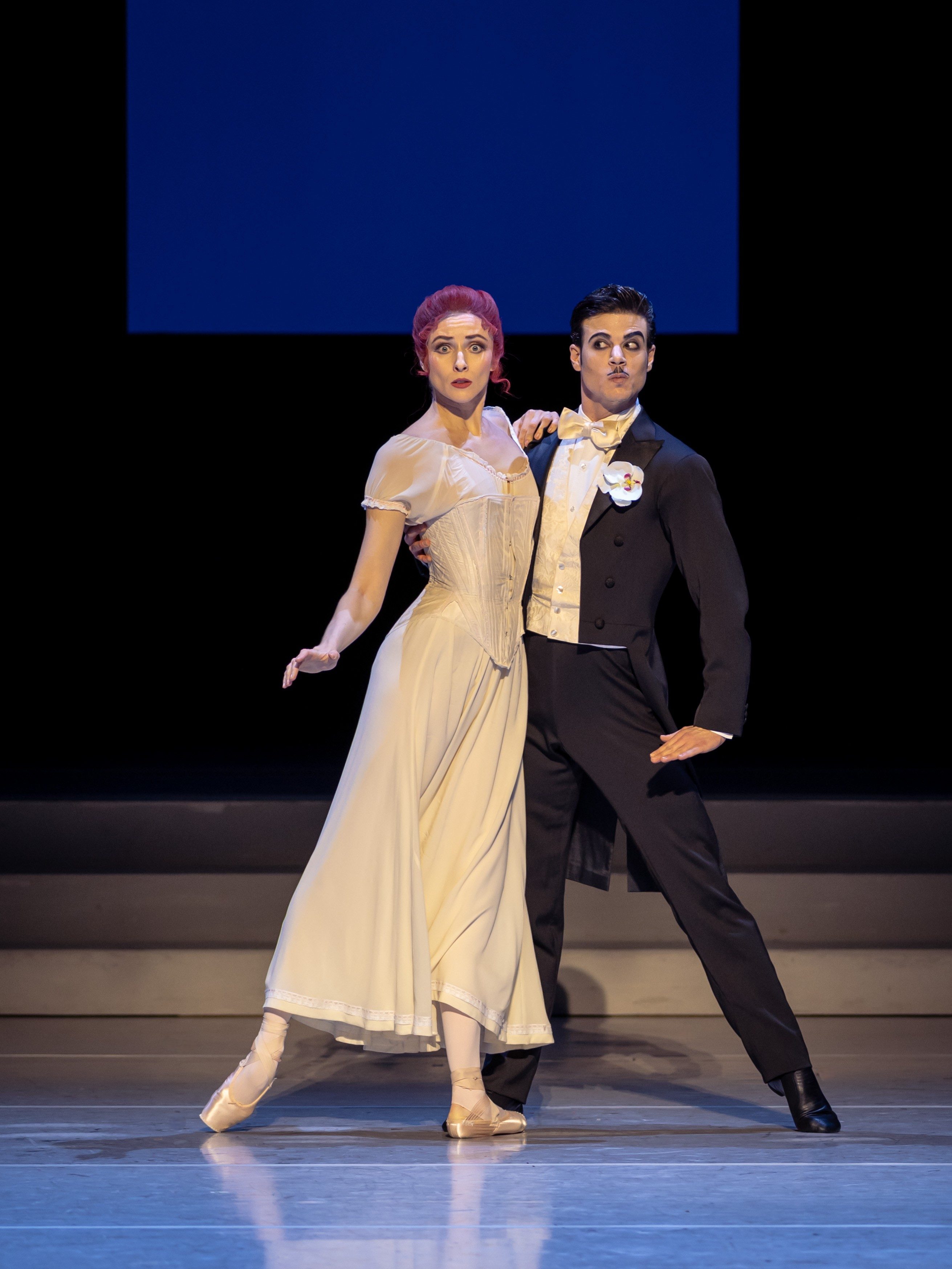 A male and female ballet dancer pose together on stage. The female dancer wears a flowing beige dress with a fitted bodice and ballet slippers, while the male dancer is dressed in formal black attire with a white shirt and bow tie. Both display expressive facial expressions.