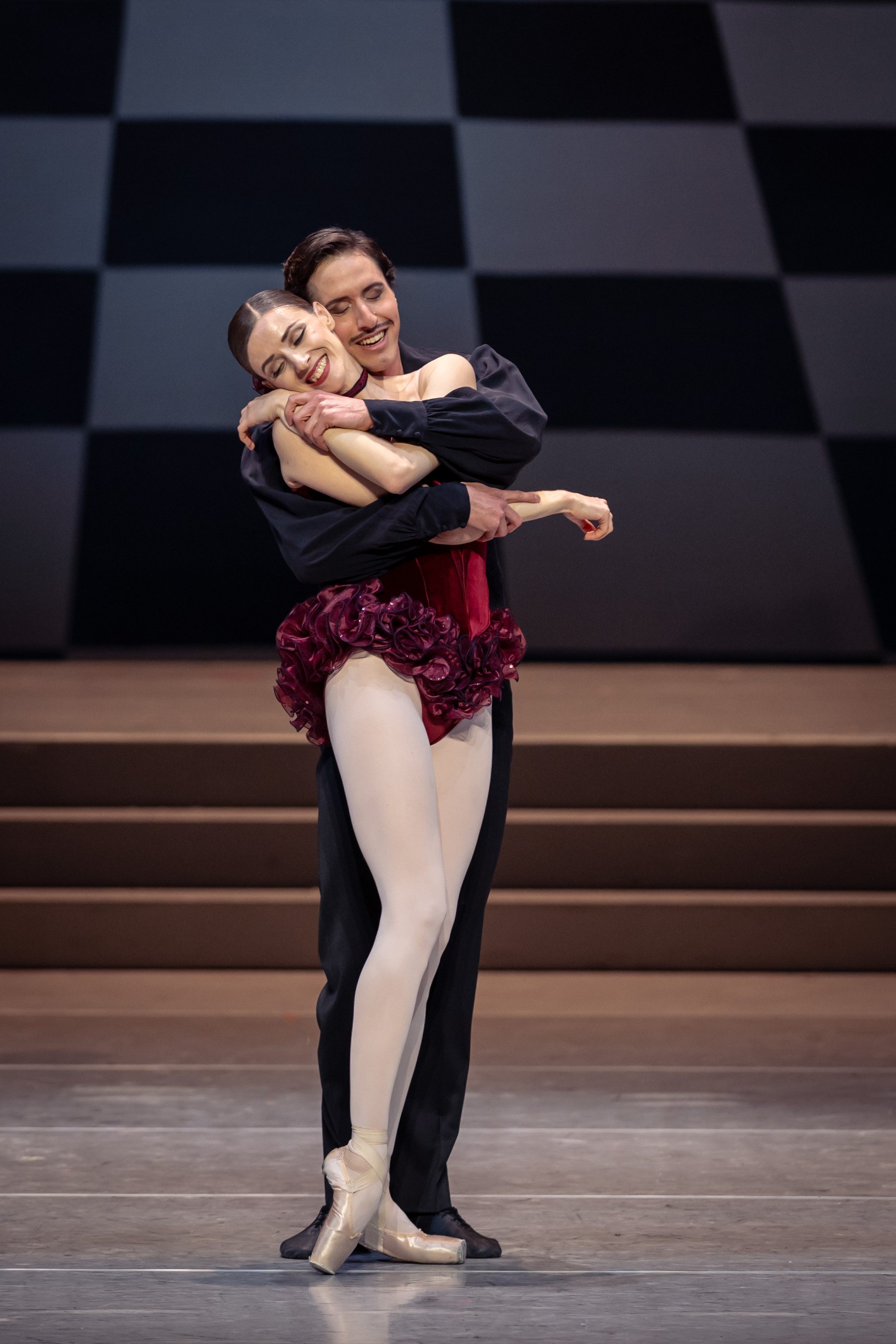 A male ballet dancer embraces a female dancer on stage. The female dancer wears a vibrant red costume with ruffles, while the male dancer is dressed in a black top. Both are smiling, conveying joy and connection amidst a neutral backdrop.