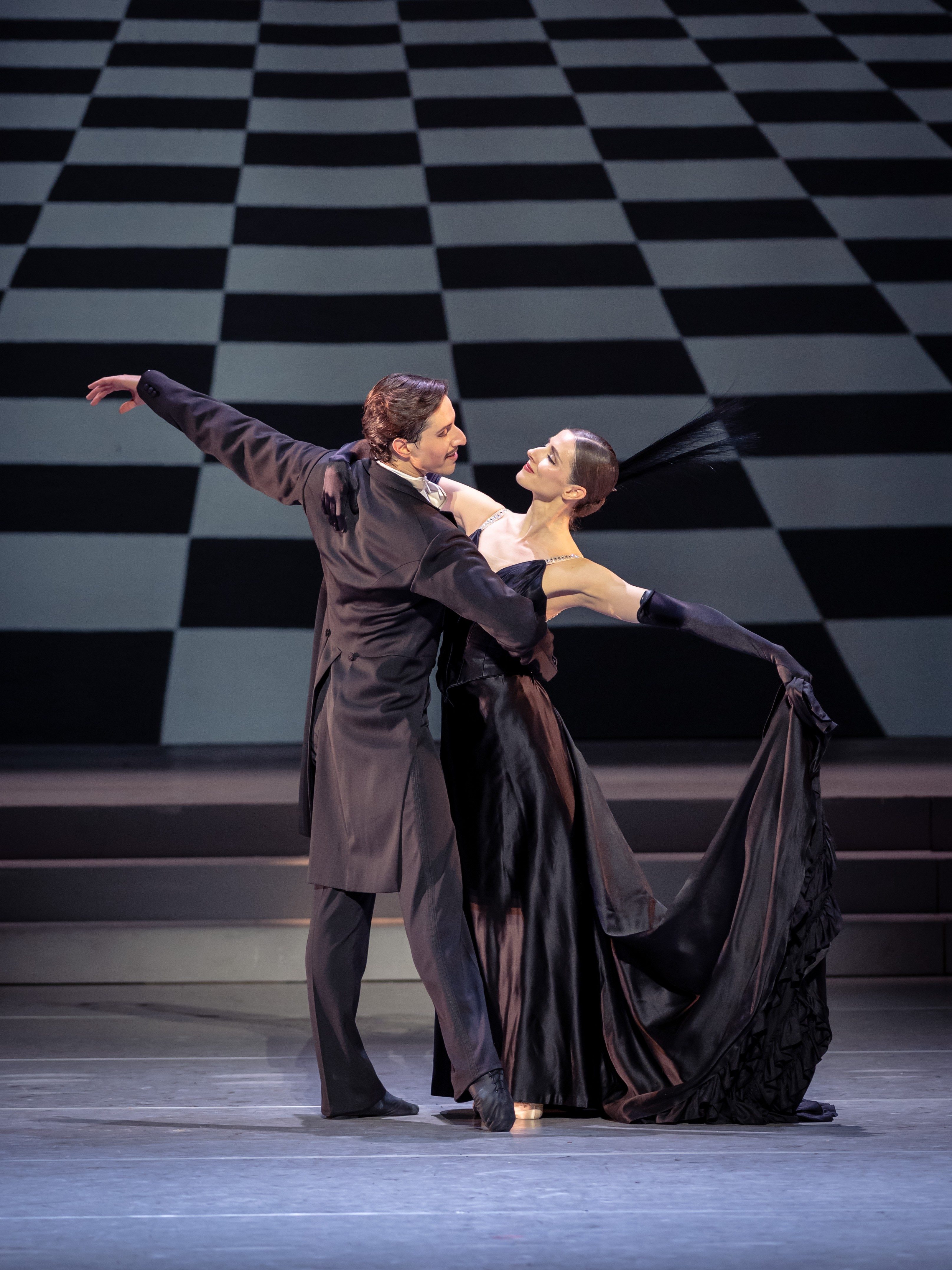 Ein Paar tanzt leidenschaftlich auf einer Bühne. Der Mann trägt einen klassischen Anzug, die Frau ein elegantes, schwarzes Abendkleid mit langen Handschuhen. Die Bewegungen sind anmutig und drücken Intimität und Harmonie aus. Hintergrund mit Schachbrettmuster, das die Dynamik des Tanzes verstärkt.