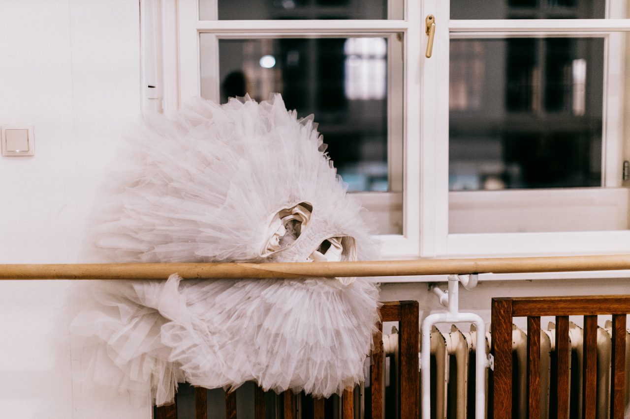 A white ballet tutu hangs on a wooden bar in a dance studio. The soft layers of tulle create a fluffy appearance, while a backdrop of a window adds a serene atmosphere to the scene. The focus remains on the tutu, showcasing its delicate fabric.