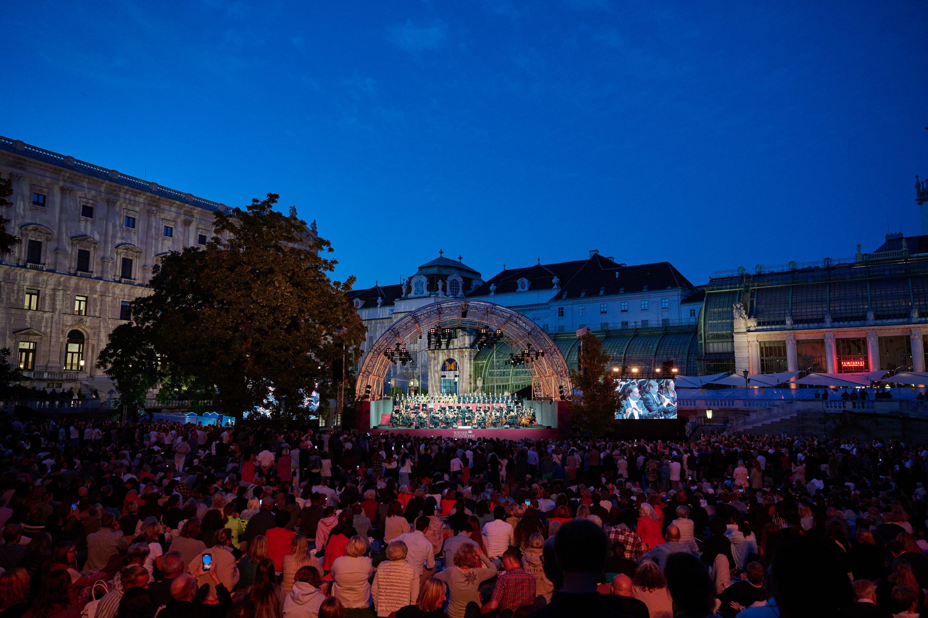 Eine große Menschenmenge sitzt auf dem Platz und schaut auf eine Bühne, die von einem Lichtbalken umrahmt ist. Im Hintergrund sind historische Gebäude zu sehen, während ein Orchester und ein Chor auf der Bühne auftreten. Es ist Abenddämmerung mit blauem Himmel.
