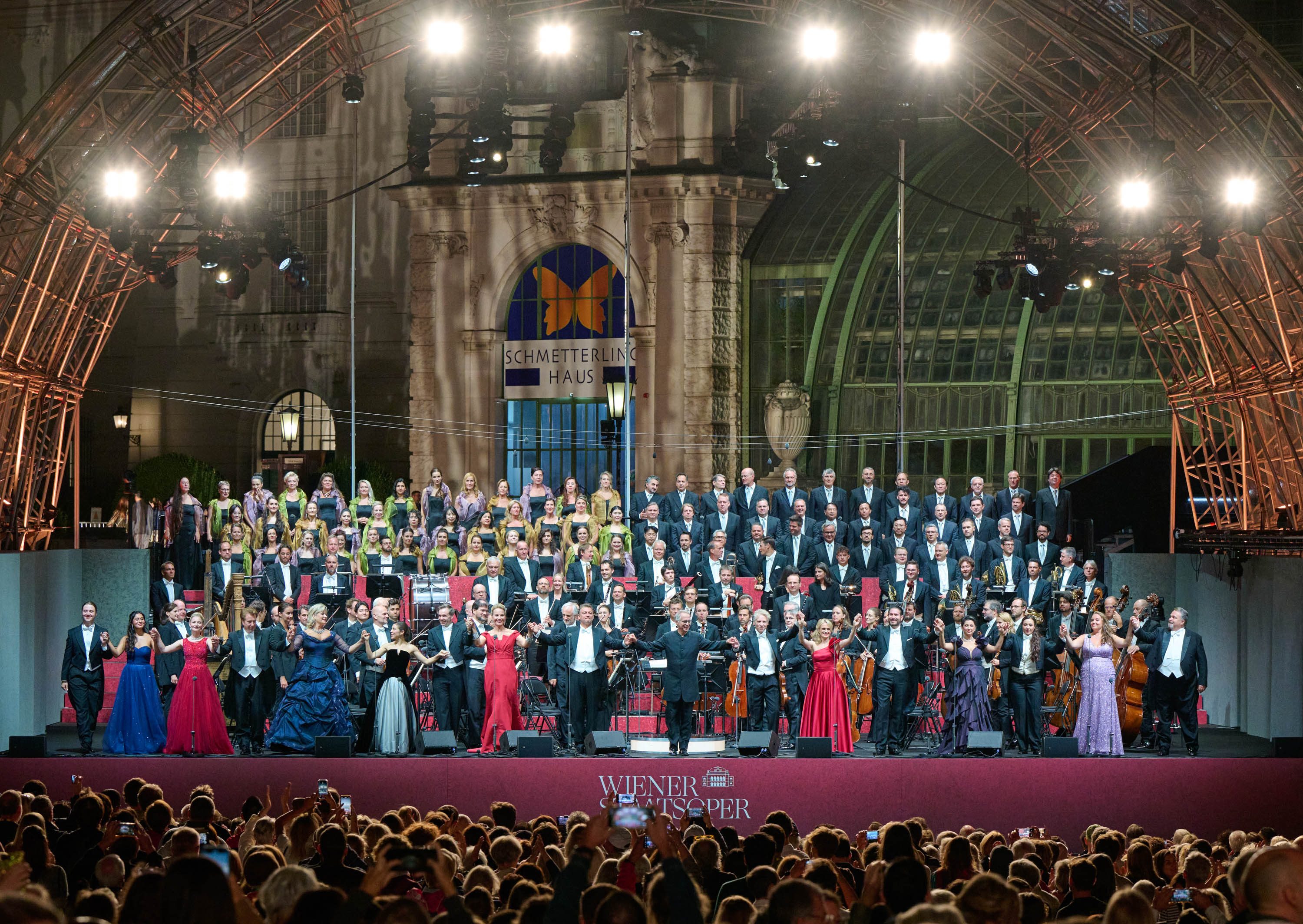 Ein großes Orchester und ein Chor treten auf einer Freiluftbühne auf. Sänger in festlicher Kleidung stehen im Vordergrund, während im Hintergrund ein Chor und eine Orchestergruppe platziert sind. Die Veranstaltung findet vor einer beleuchteten Kulisse des Schmetterlingshauses statt und zieht eine große Menschenmenge an.