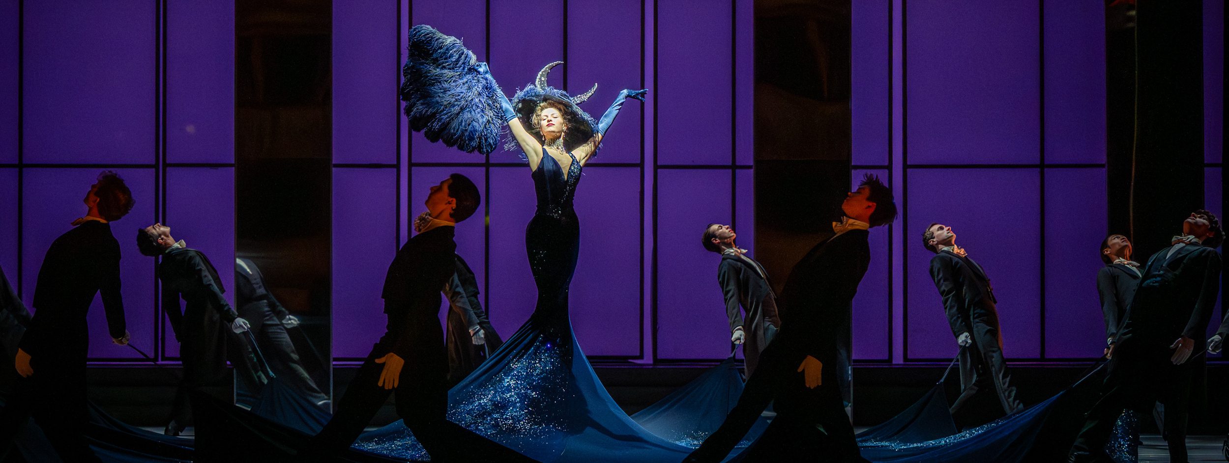 Eine dramatische Bühnenszene mit einer Sängerin in einem glitzernden, schwarzen Kleid und großen, feenhaften Flügeln. Sie hebt die Arme in einer kraftvollen Pose, umgeben von mehreren Tänzern in eleganter Abendkleidung, die sich zur Seite neigen und eine hypnotisierende Atmosphäre schaffen.