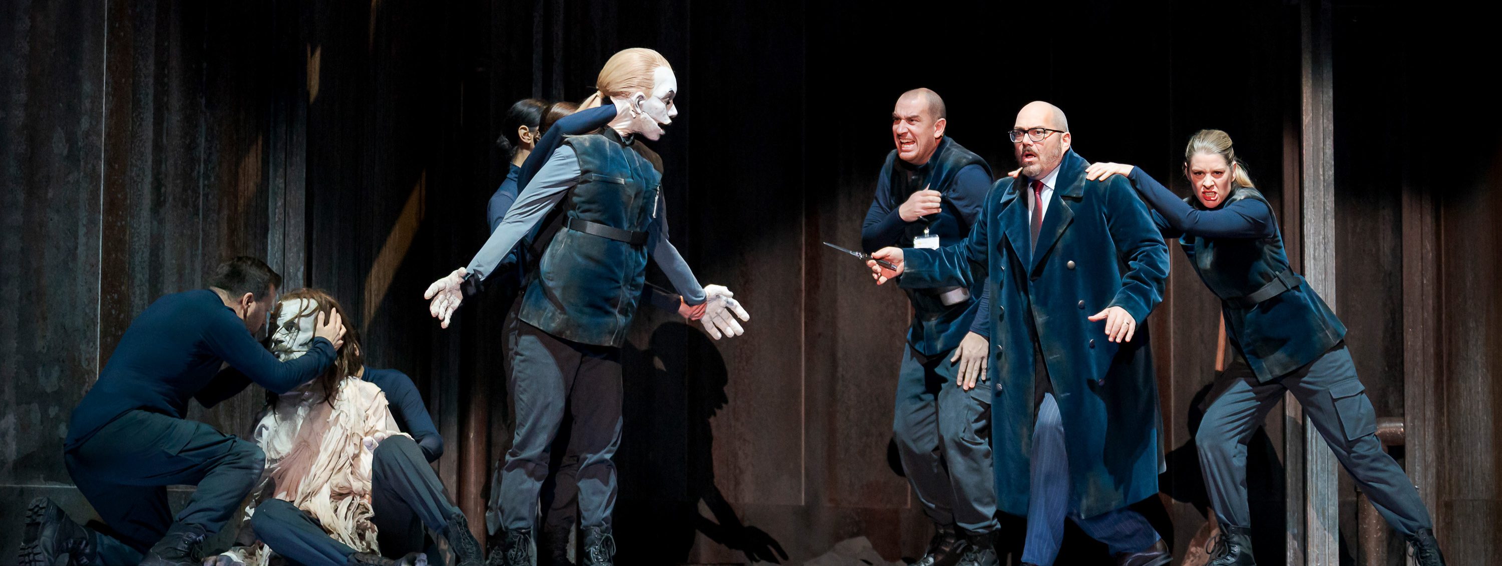 Eine dramatische Theateraufführung zeigt eine Gruppe von Personen in Uniformen, die emotional reagieren. Eine Person kniet vor einer gesichtslosen Figur, während andere in Aufregung miteinander sprechen. Der Hintergrund besteht aus dunklem Holz und verleiht der Szene eine verstörende Atmosphäre.