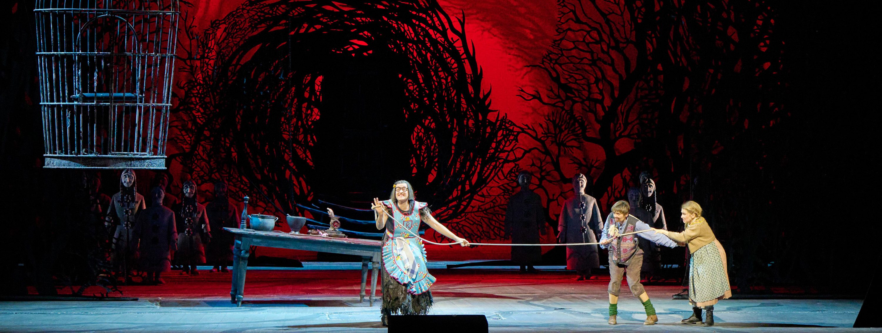 The image depicts a dramatic stage scene with three characters interacting on a set. One character, in a colorful outfit, is holding a rope connected to another, while a third character stands nearby. The background features abstract, dark designs, adding to the overall theatrical atmosphere.