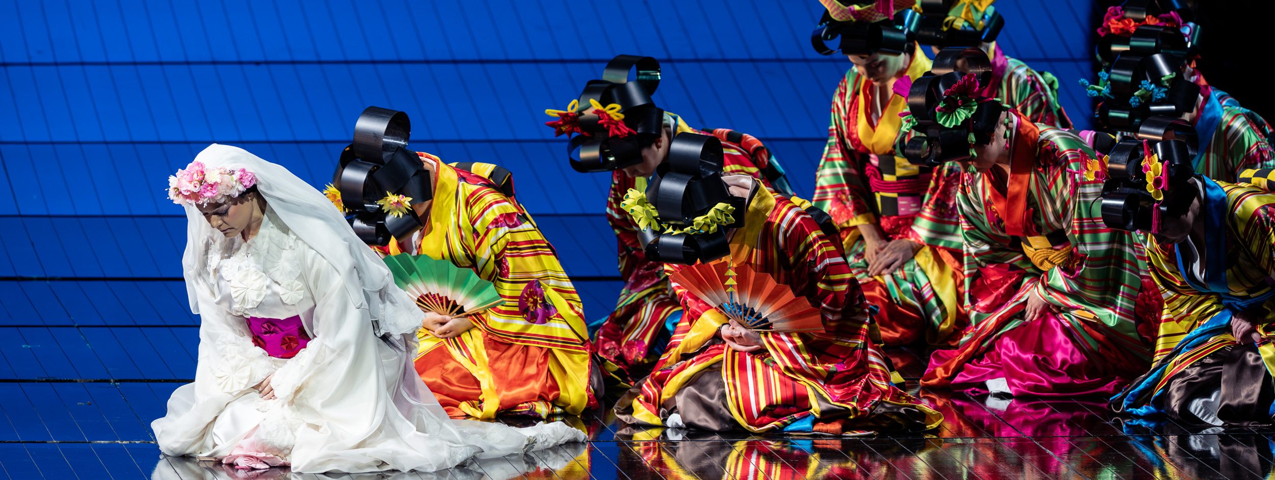 Eine Frau in einem weißen traditionellen Kleid mit Blumenschmuck auf dem Kopf kniet auf dem Boden. Um sie herum sitzen weitere Frauen in bunten, gestreiften Gewändern, die ihre Köpfe geneigt haben und Fächer in ihren Händen halten. Der Hintergrund ist blau.