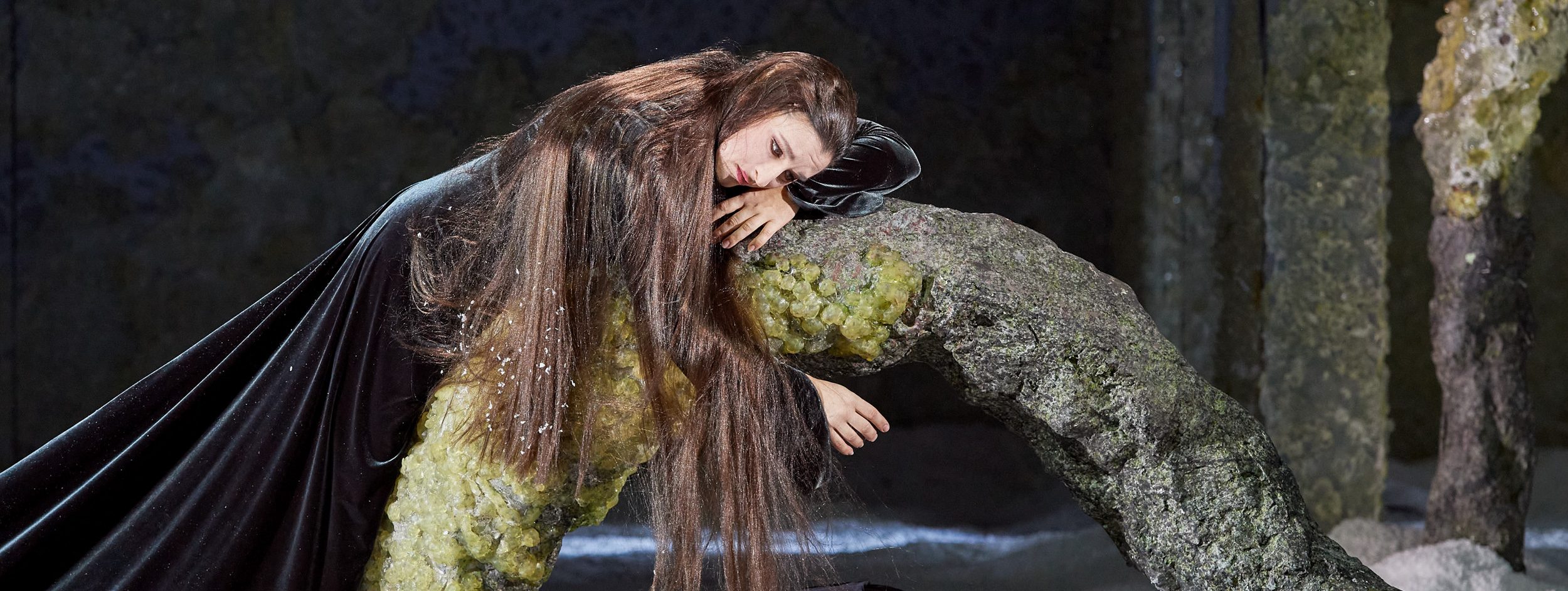 Eine Person mit langen, fließenden Haaren liegt auf einem geformten, steinähnlichen Objekt. Sie trägt ein schwarzes Gewand und hat einen nachdenklichen Ausdruck, während sie auf dem Objekt ruht, das mit grünen Kristallen bedeckt ist. Der Hintergrund ist dunkel und mystisch.