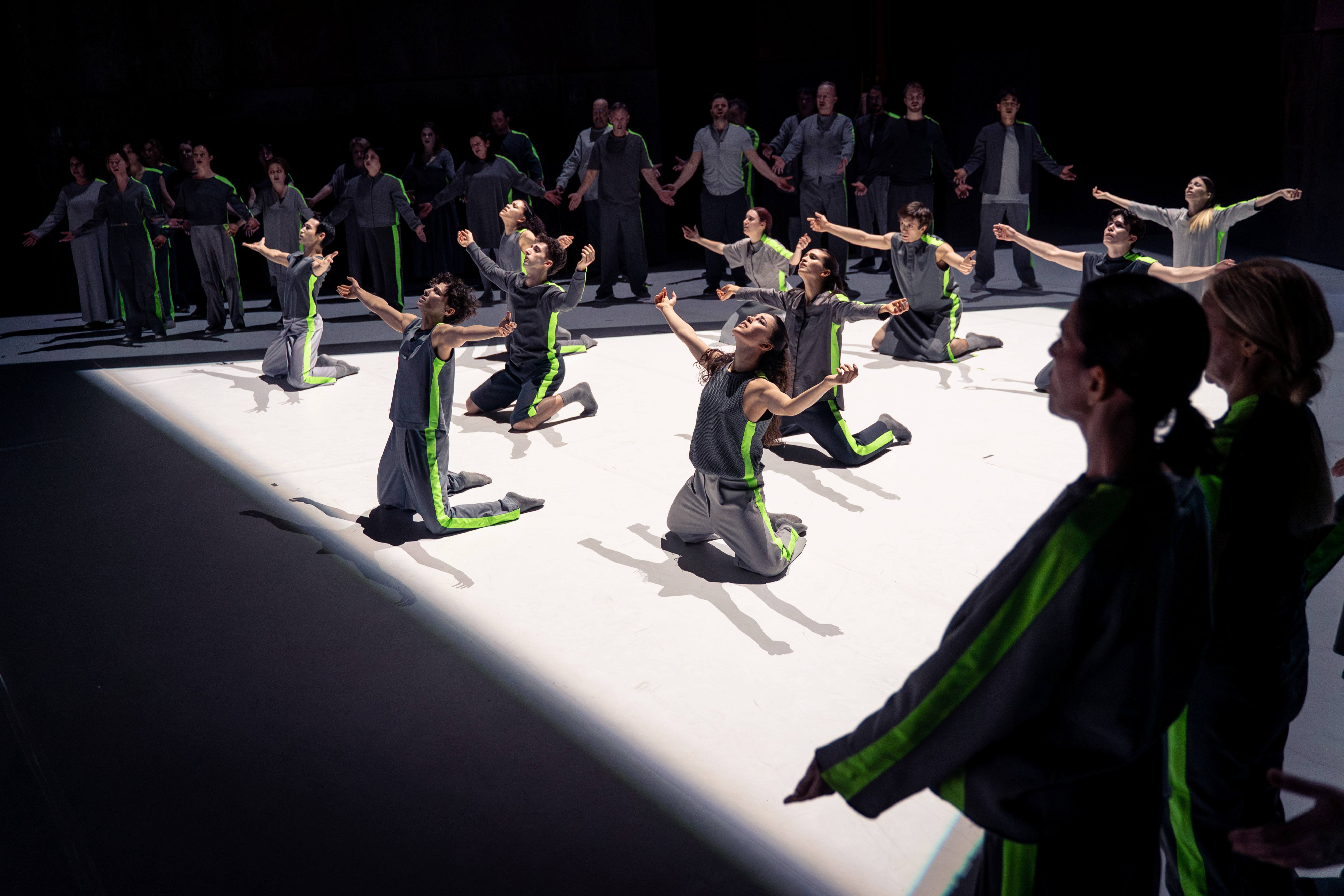 Eine Gruppe von Tänzern in grauen Outfits mit grünen Akzenten führt eine ausdrucksstarke Choreografie auf einer hellen Bühne auf. Einige Tänzer knien, während andere ihre Arme zur Decke heben. Die Szene vermittelt Gefühle von Hingabe und Gemeinschaft.