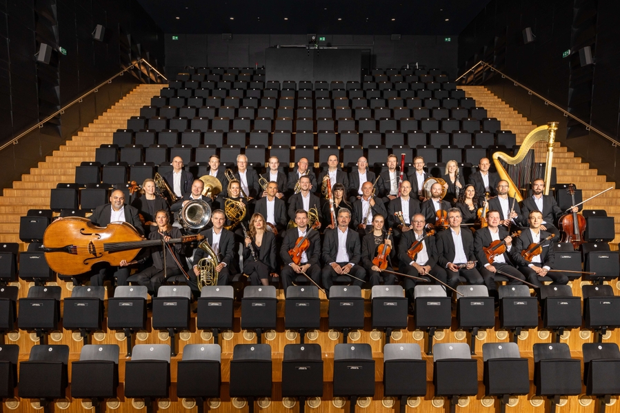 A diverse orchestra poses in a modern auditorium. They are seated in multiple rows, surrounded by empty black chairs. Musicians hold various instruments, including strings, brass, and woodwinds, while dressed in formal black attire, creating a sense of unity and professionalism.