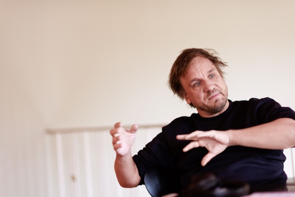 Ein Mann mit ungepflegtem Haar in einem dunklen Pullover sitzt auf einem Stuhl. Er gestikuliert mit seinen Händen und spricht möglicherweise leidenschaftlich über ein Thema. Der Hintergrund ist schlicht und unauffällig, was den Fokus auf den Mann lenkt.