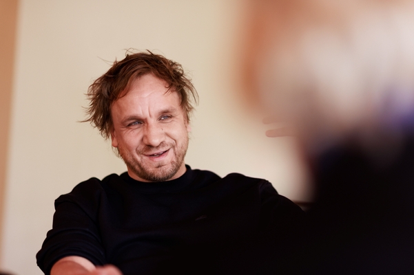 Ein lächelnder Mann mit wuscheligem Haar trägt ein schwarzes Oberteil. Er sitzt in einem Gespräch und hat einen freundlichen Ausdruck im Gesicht. Im Hintergrund ist eine leicht verschwommene Person zu erkennen, die gestikuliert. Die Umgebung wirkt hell und einladend.