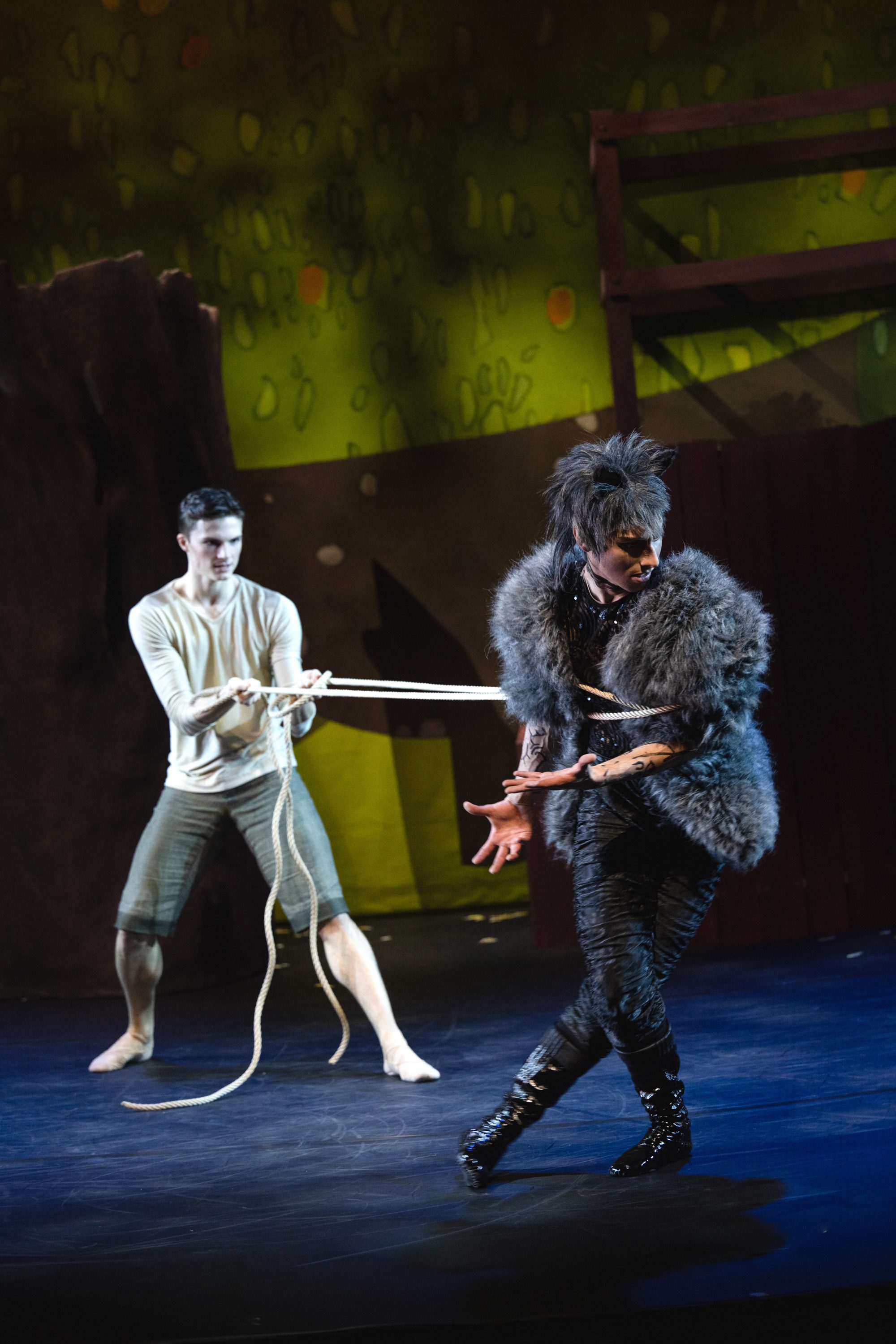 Zwei Tänzer auf einer Bühne: Ein Mann zieht mit einem Seil einen anderen, der mit einem auffälligen Pelzkleidungsstück bekleidet ist. Der Hintergrund zeigt eine künstlerische Darstellung von Pflanzen und Bäumen, die eine lebendige Bühnenatmosphäre schaffen.