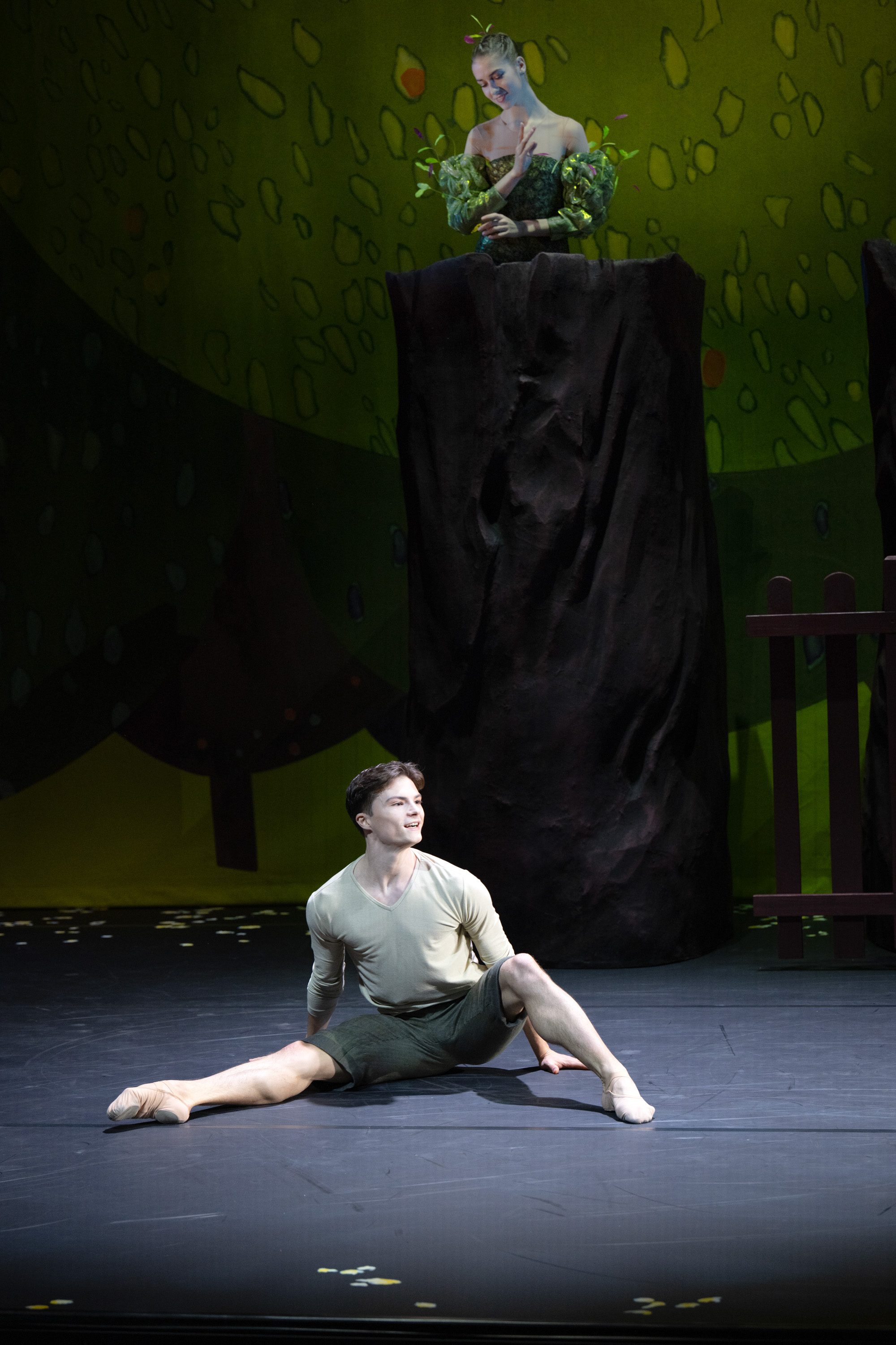 Ein Tänzer in schlichten Kostümen sitzt auf der Bühne und führt eine elegante Pose aus. Im Hintergrund steht eine weitere Tänzerin, die in einem Pflanzenkostüm gekleidet ist. Die Szenerie ist von einer bunten, natürlichen Kulisse umgeben, die eine märchenhafte Atmosphäre schafft.
