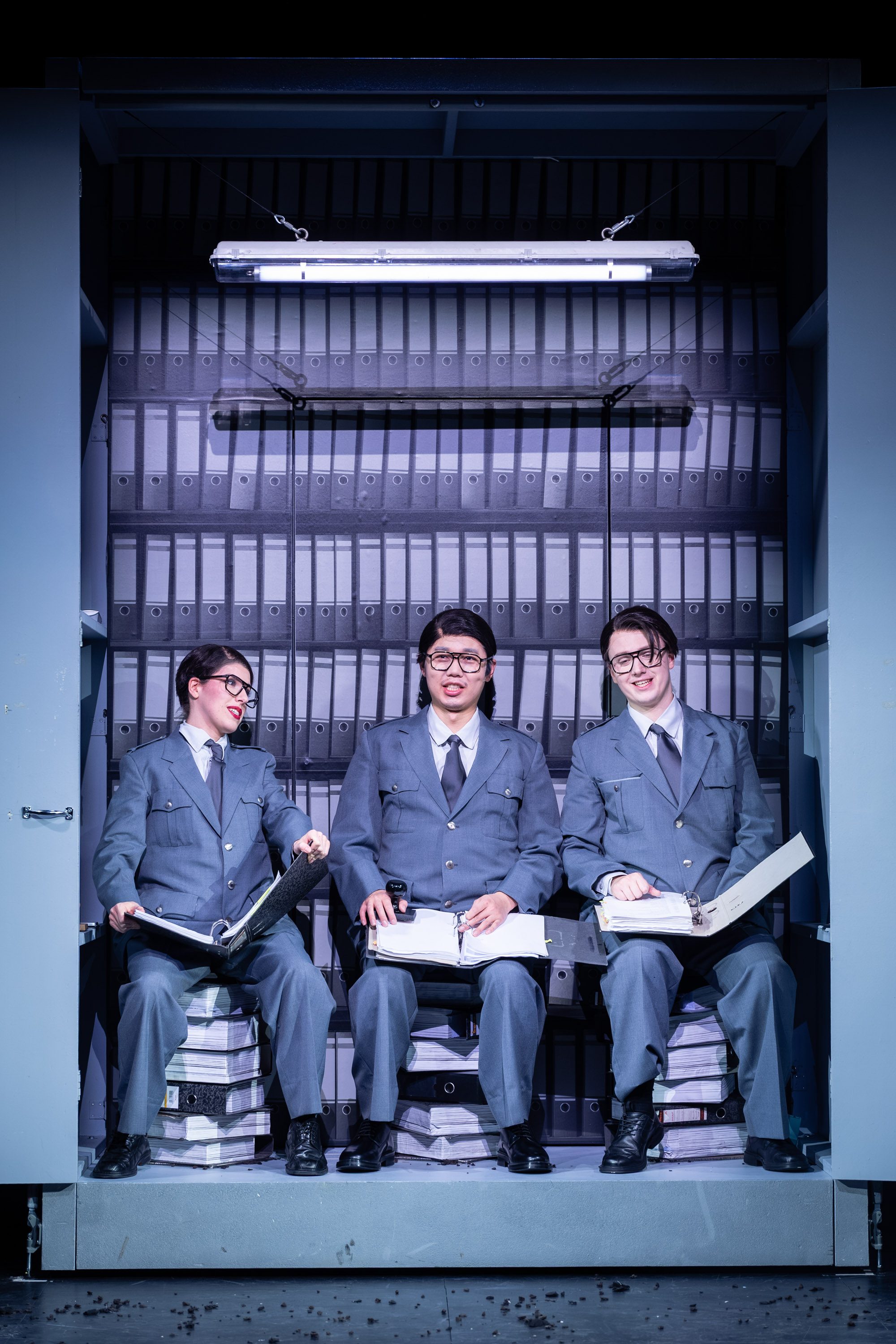 Drei Personen in grauen Anzügen sitzen auf gestapelten Aktenordnern. Sie halten Akten in den Händen und schauen in die Kamera, während eine Deckenlampe den Raum beleuchtet. Im Hintergrund sind Regale mit Aktenordnern sichtbar.