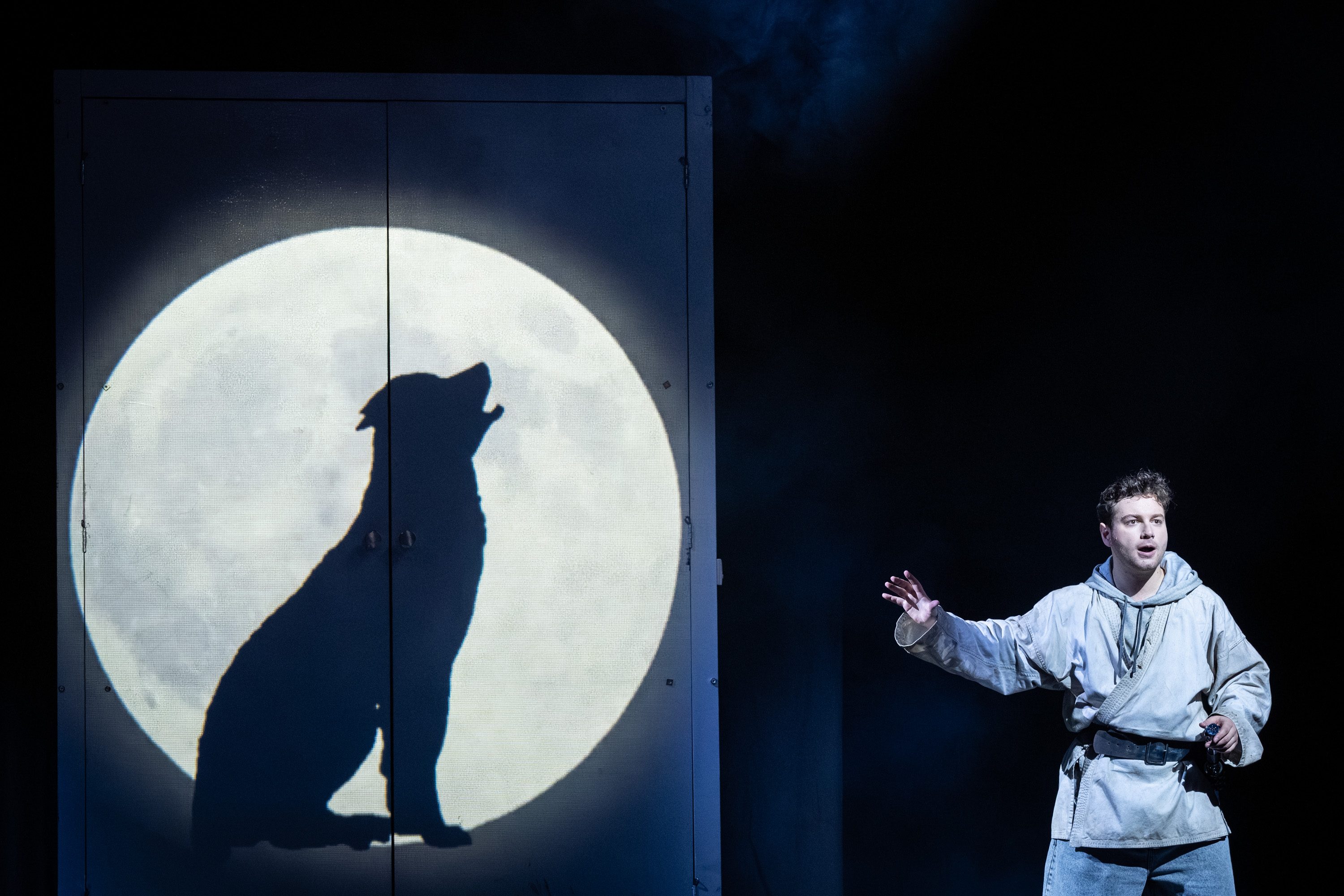 Ein Schauspieler steht in einem hell erleuchteten Raum und hebt eine Hand. Hinter ihm projiziert der Mond den Schatten eines heulenden Hundes auf eine Wand. Die Szene vermittelt eine dramatische Stimmung.