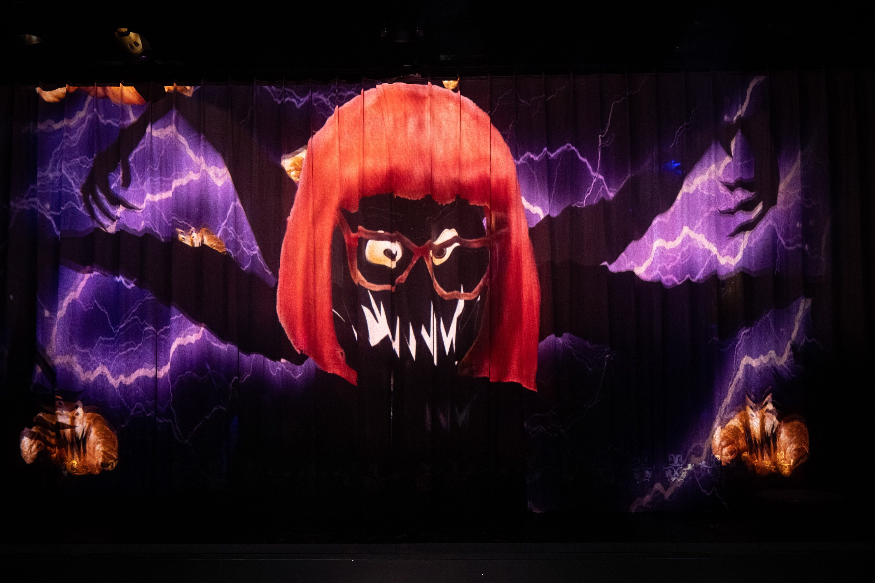 Eine grafische Darstellung zeigt ein groteskes Gesicht mit roten Haaren und Brille, umgeben von dunklen Armen. Der Hintergrund ist mit lila Blitzen und schemenhaften Tieren gestaltet, was eine düstere Atmosphäre erzeugt.