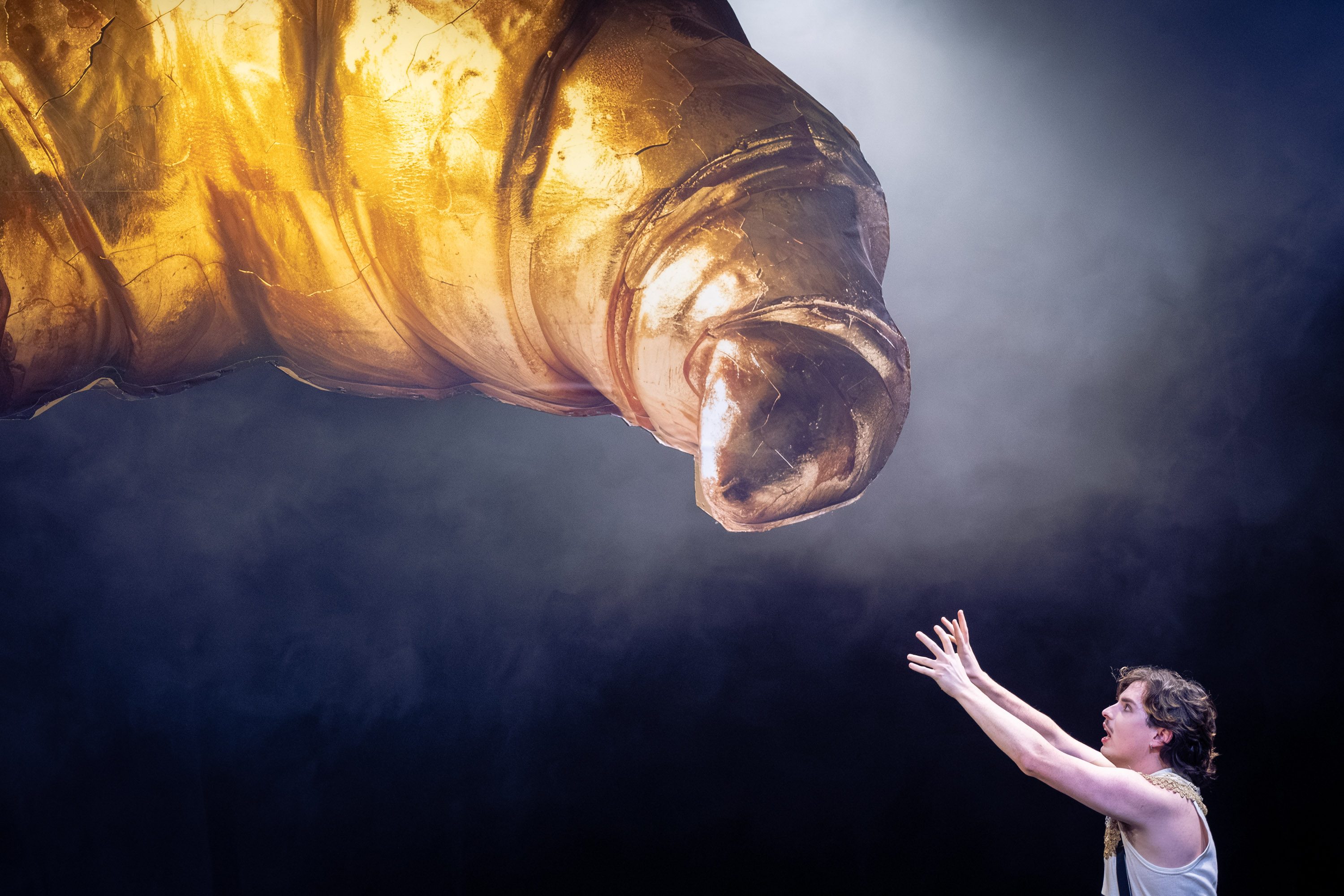 Ein Künstler steht auf der Bühne und streckt seine Hände nach einem großen, goldenen Finger aus, der von oben auf ihn herabzeigt. Der Hintergrund ist dunkel und neblig, was die dramatische Atmosphäre verstärkt.