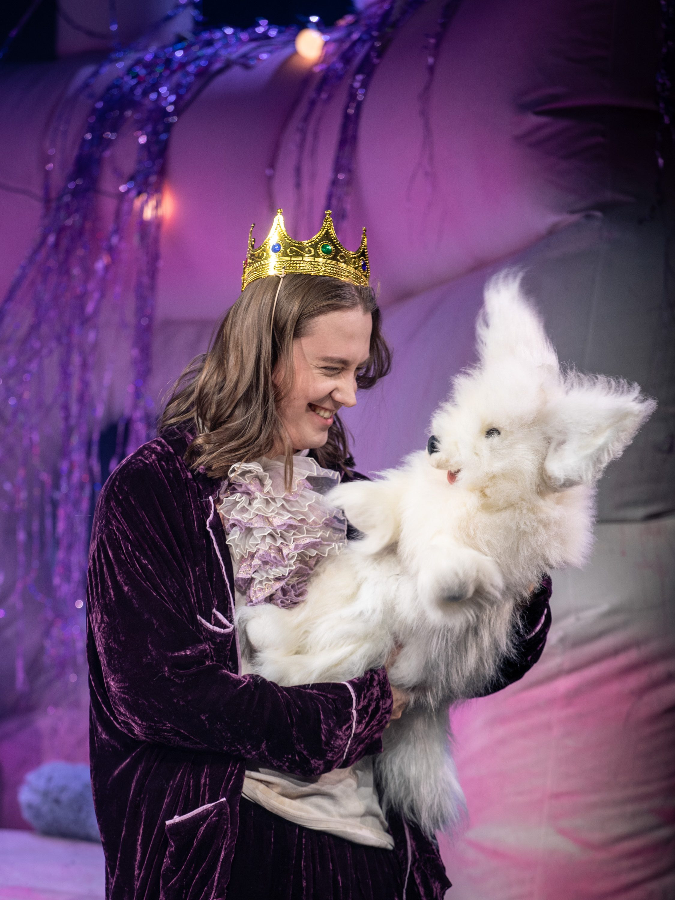 Eine Person mit einer goldenen Krone lächelt und hält ein flauschiges, weißes Stofftier in der Hand. Die Szenerie hat einen farbenfrohen, festlichen Hintergrund, der eine spielerische Atmosphäre vermittelt.