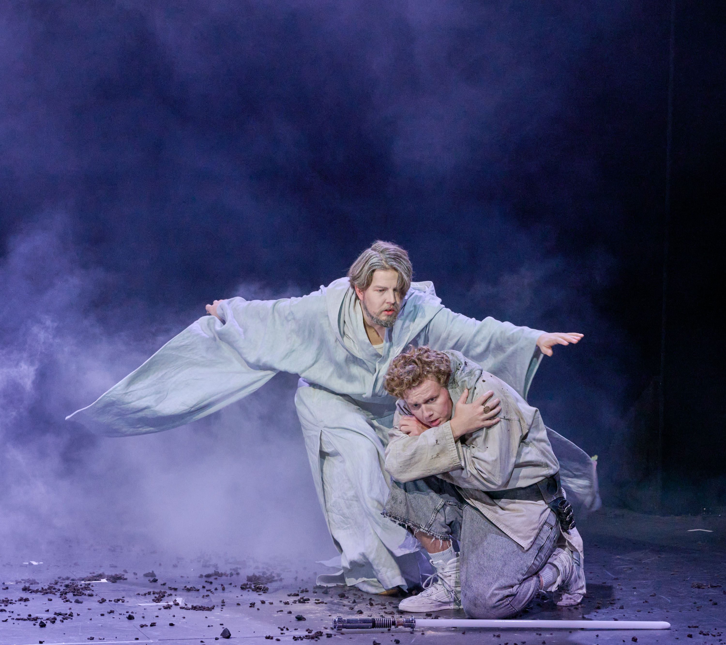 Zwei Schauspieler auf einer Bühne, umgeben von Nebel. Der erste, in einem grauen Gewand, steht schützend hinter dem zweiten, der kniet und sichtlich verängstigt wirkt. Die Szene vermittelt Gefühle von Angst und Schutz in einer dramatischen Atmosphäre.