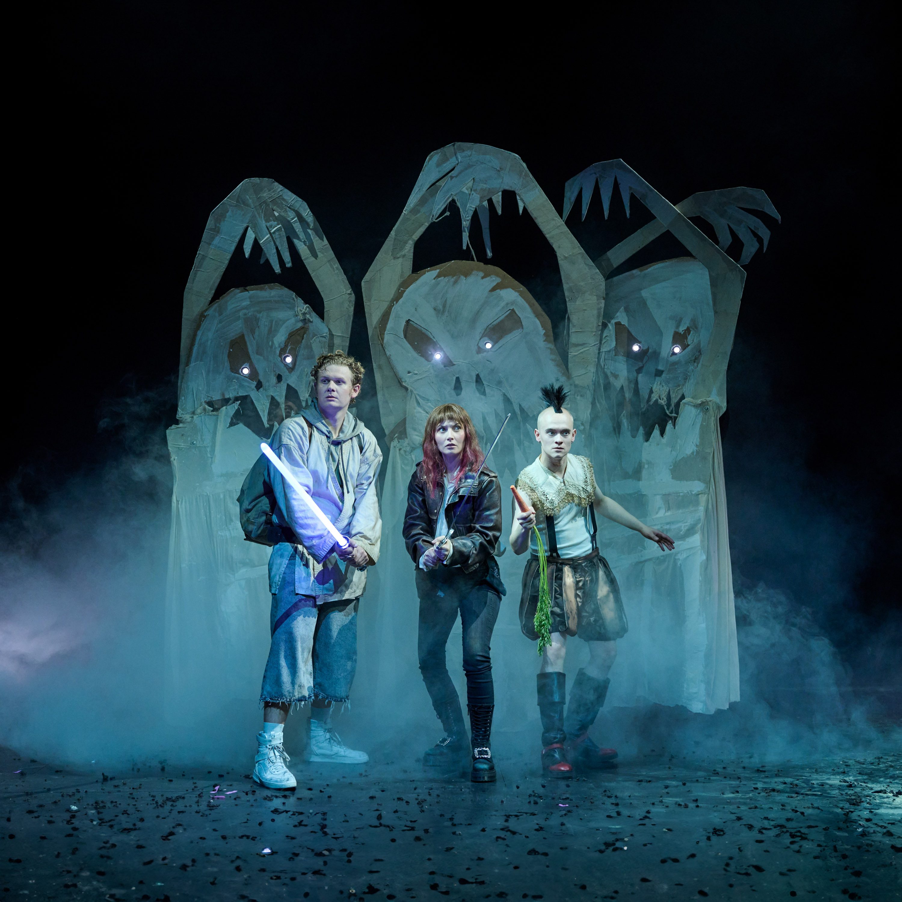 Drei Charaktere stehen vor einem gruseligen Hintergrund aus Papierfiguren, die gesichtslos und bedrohlich wirken. Einer hält ein leuchtendes Schwert, die andere eine Art Stock, während der dritte eine Karotte in der Hand hält. Rauch umhüllt die Szene, die eine spannende Atmosphäre erzeugt.