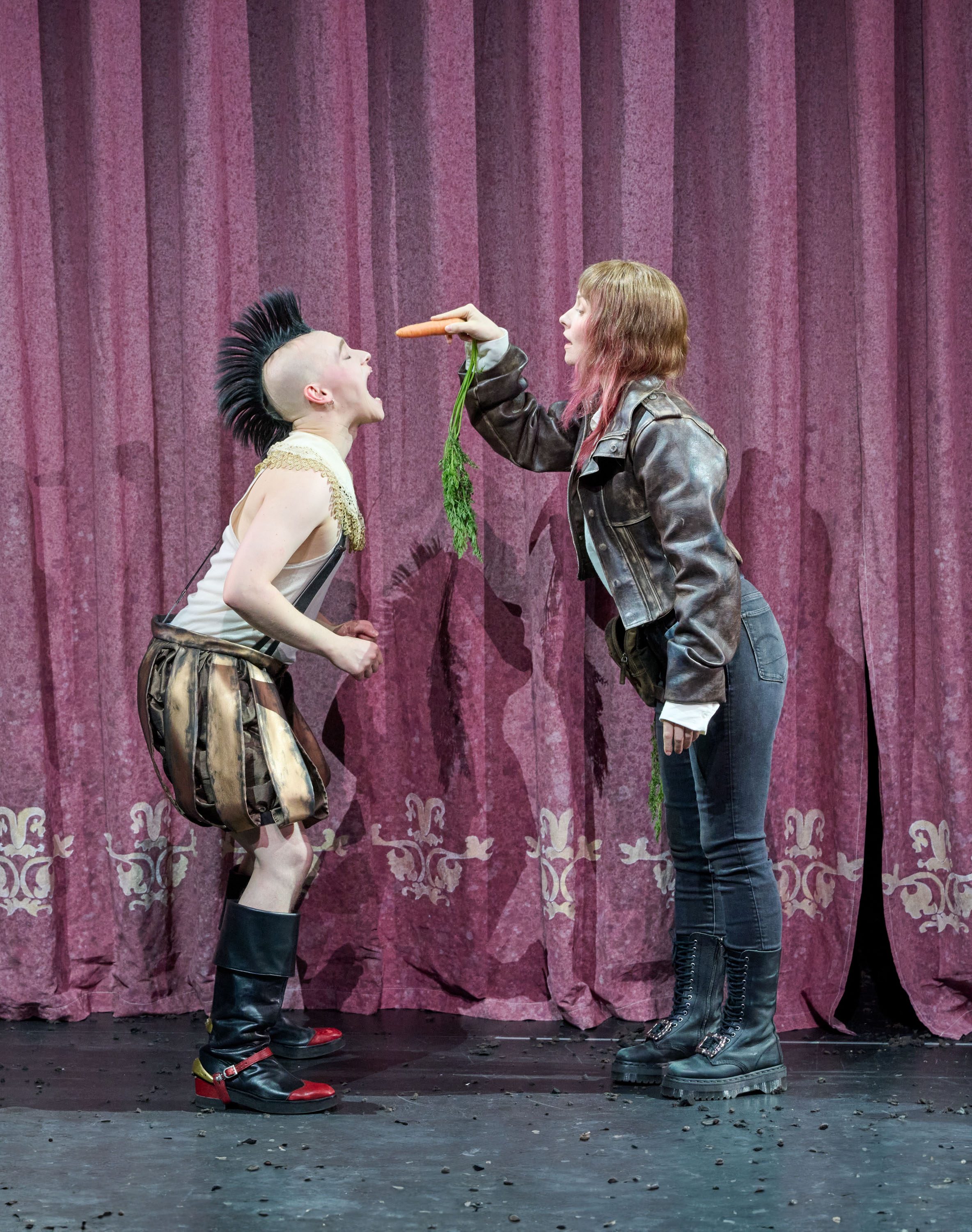 Auf der Bühne steht eine Person mit Mohawk-Frisur, die laut schreit, während eine andere Person mit langen Haaren eine Karotte in Richtung ihres Mundes hält. Der Hintergrund besteht aus roten Vorhängen. Die Szene vermittelt eine angespannte, aber humorvolle Interaktion.
