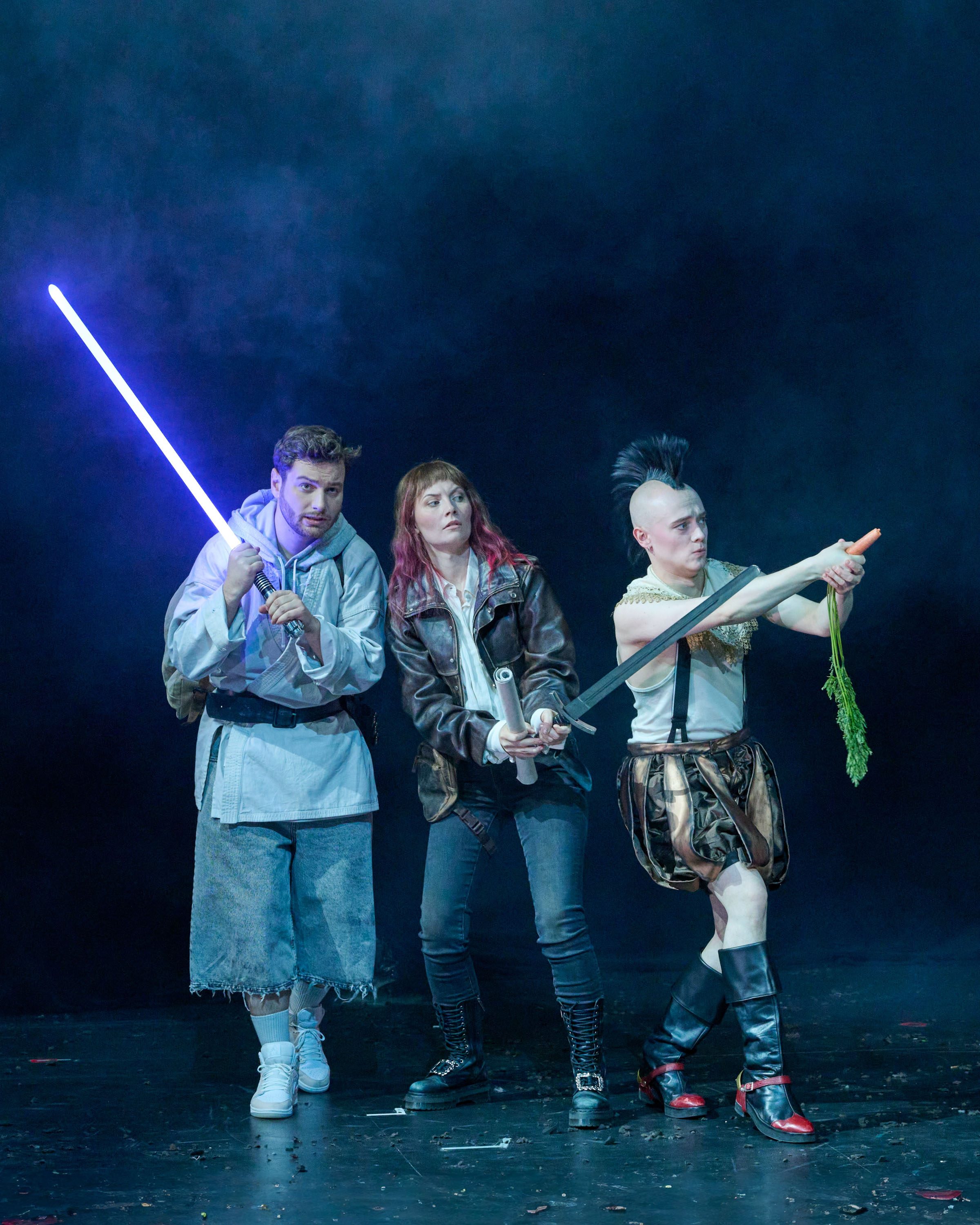 Drei Schauspieler stehen auf einer dunklen Bühne. Sie tragen fantasievolle Kostüme und halten verschiedene Requisiten: ein Lichtschwert, eine zusammenklappbare Pistole und eine Karotte. Ihre Posen deuten auf eine dramatische Szene hin, während sie in die Ferne blicken.