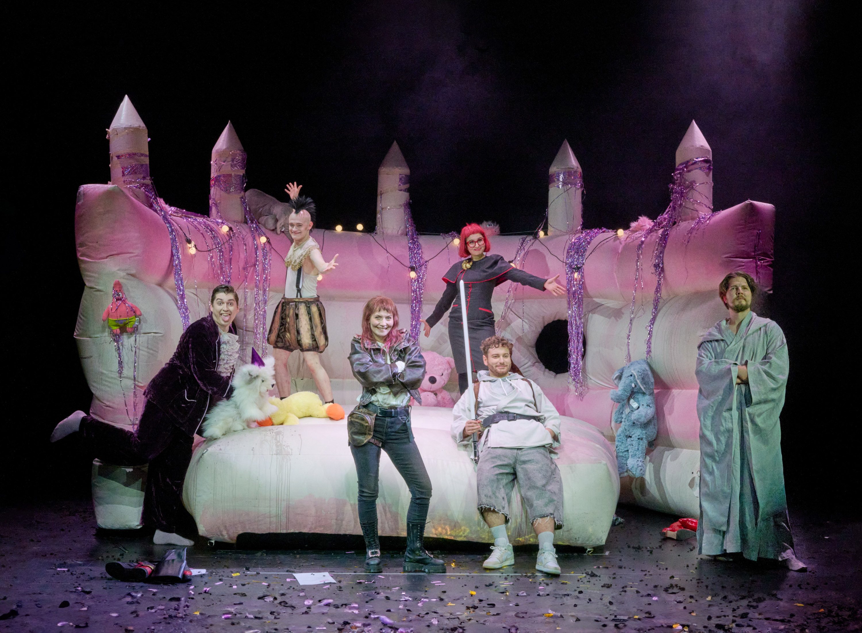Eine Gruppe von sechs Personen posiert auf einer großen, bunten Hüpfburg. Sie tragen verschiedene, ausgefallene Kostüme und zeigen fröhliche Gesten. Die Umgebung ist mit Verzierungen und Konfetti geschmückt, was eine festliche Atmosphäre schafft.