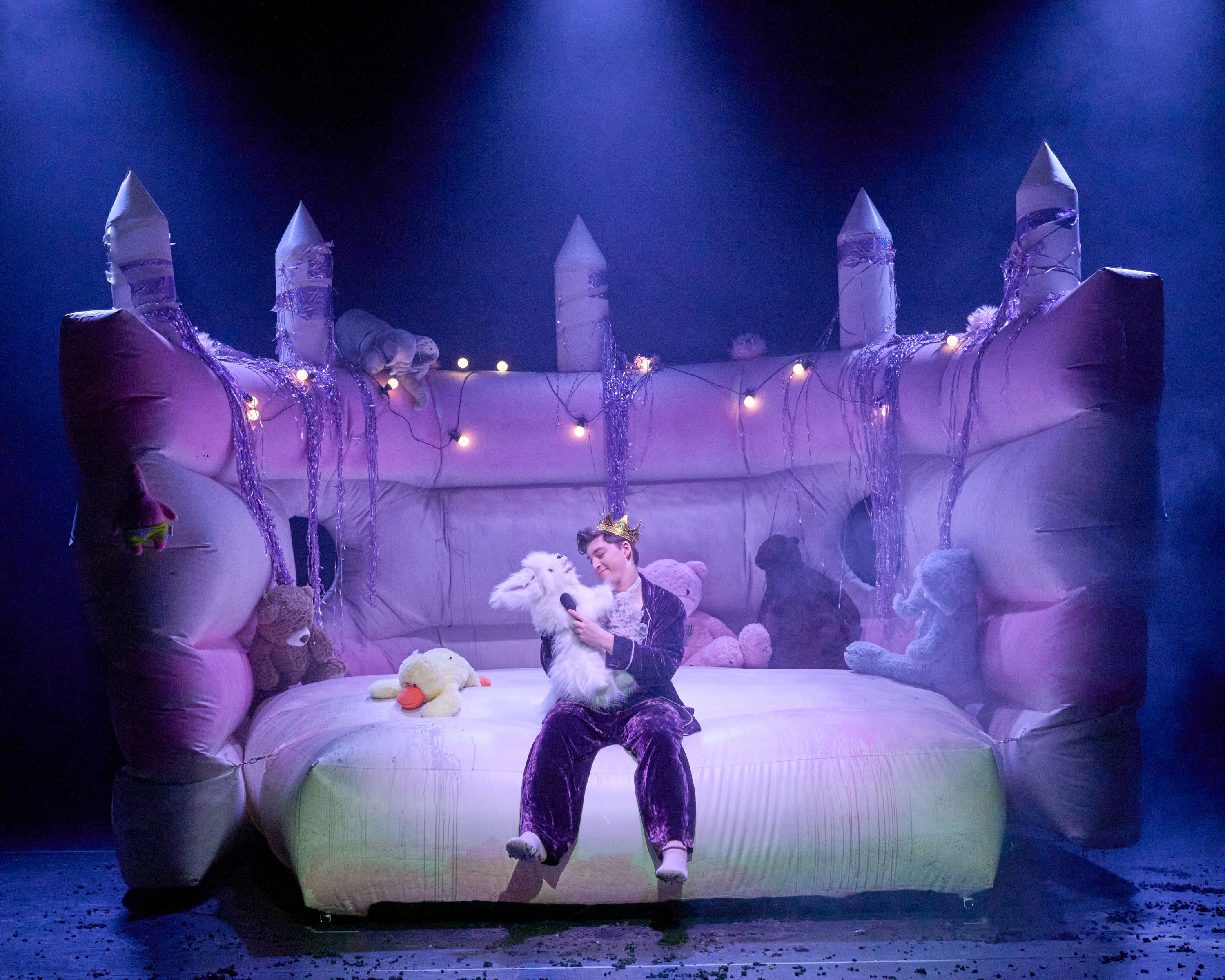 Ein junger Mensch mit einer Krone sitzt auf einer großen, aufblasbaren Matratze, umgeben von Kuscheltieren. Sie halten ein weißes Plüschtier und sprechen mit einem Mikrofon. Sanftes Licht und eine märchenhafte Atmosphäre dominieren die Szene.