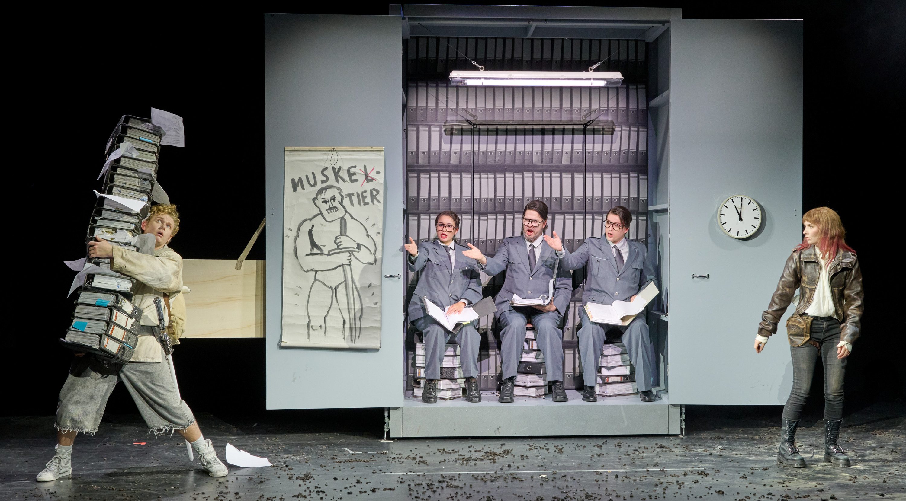 Auf der Bühne steht eine Szene mit vier Personen. Eine Person trägt einen großen Stapel Akten, während drei andere in Anzügen auf Kisten sitzen und gestikulierend diskutieren. Neben ihnen hängt ein Plakat mit einer Zeichnung und dem Wort „MUSKELTIER“. In der Ecke ist eine Wanduhr sichtbar.