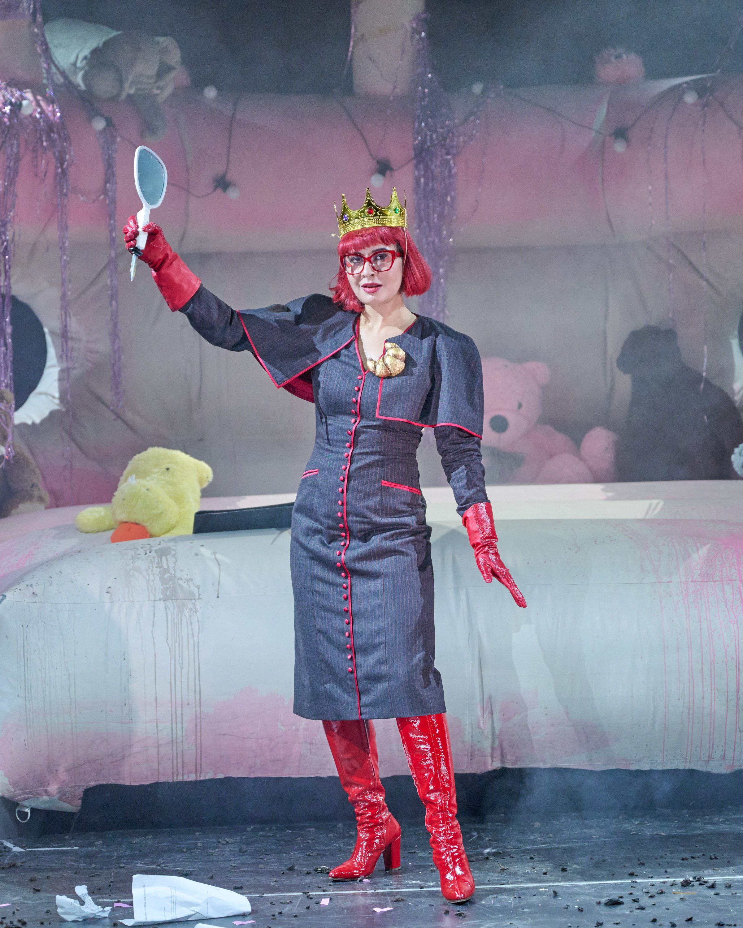 Eine Person in einer auffälligen, königlichen Kleidung hält einen Handspiegel hoch. Sie hat rote Haare, eine Krone und rote Handschuhe an. Im Hintergrund sind verschwommene Umrisse von Plüschtieren und eine dekorierte Kulisse zu sehen, um eine fantasievolle Atmosphäre zu schaffen.