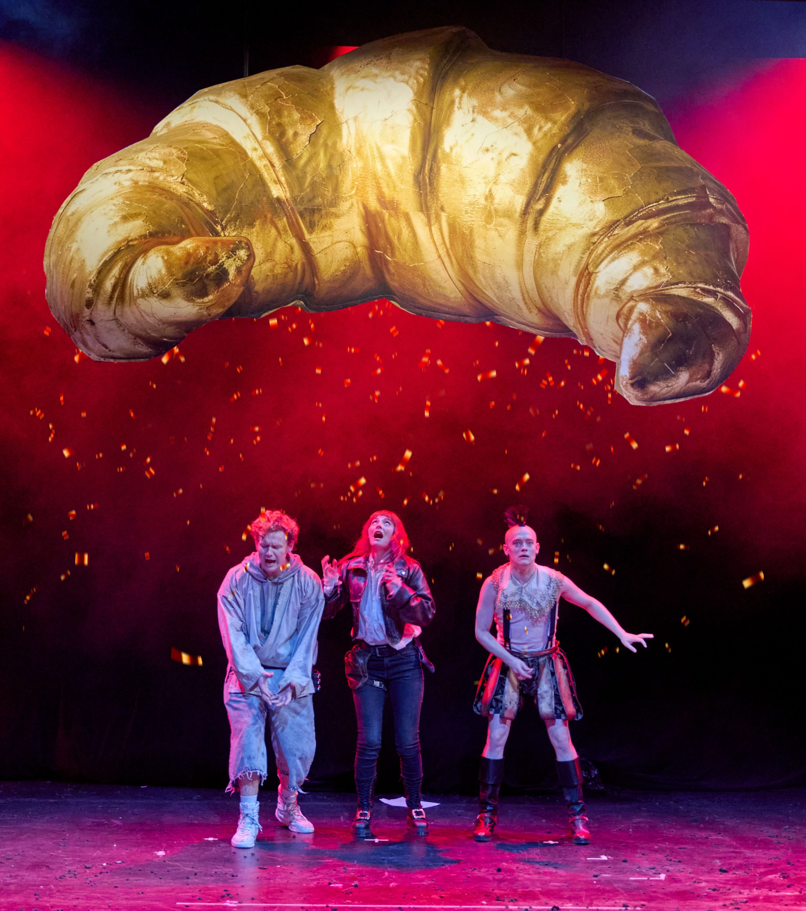 Drei Schauspieler stehen auf einer Bühne, während ein riesiges, goldenes Croissant über ihnen schwebt. Die Figuren zeigen verschiedene Emotionen: Schreck und Überraschung. Im Hintergrund leuchtet rotes Licht, das eine dramatische Stimmung erzeugt.