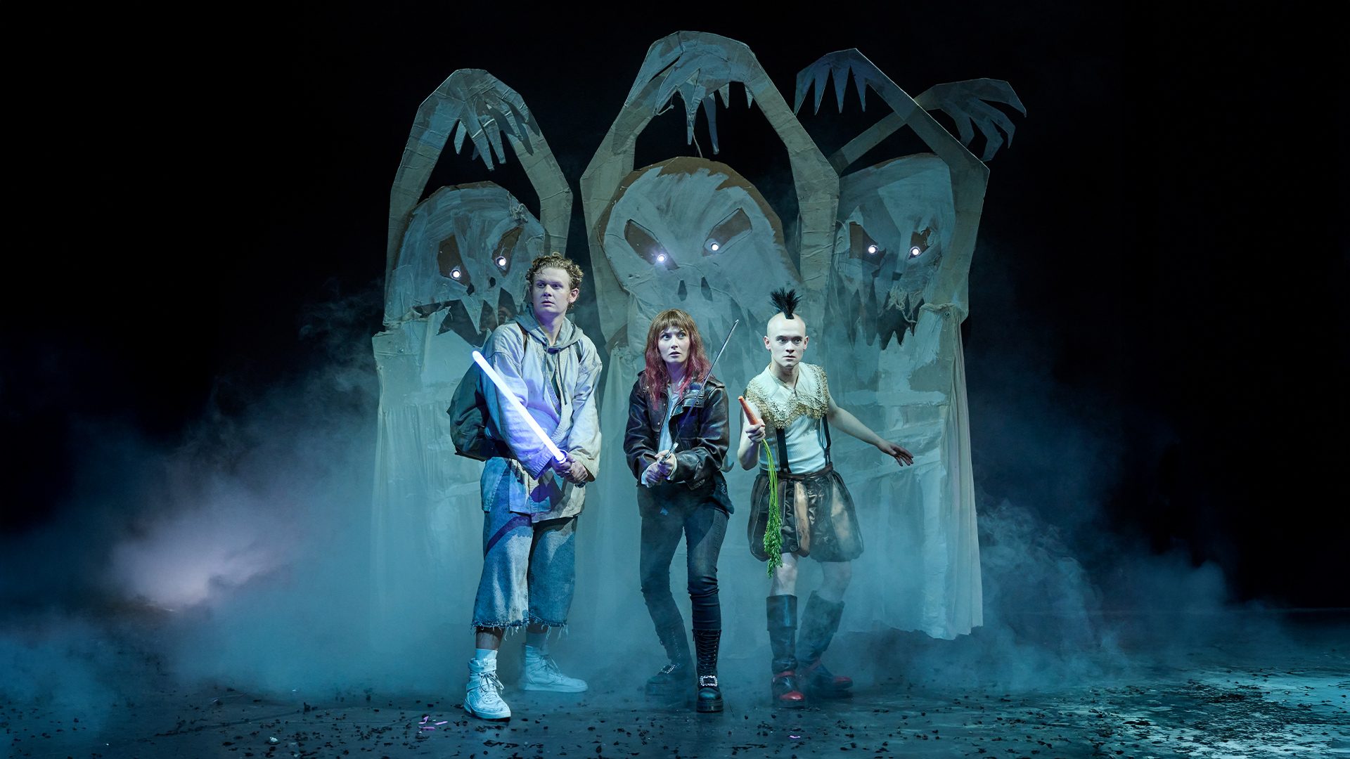 Drei Schauspieler stehen im Nebel auf einer Bühne, umgeben von bedrohlichen Pappfiguren mit gesichtslosen Masken. Sie halten unterschiedliche Requisiten: ein Lichtschwert, ein Gewehr und eine Karotte. Die Szenerie vermittelt eine spannende, fantasievolle Stimmung.