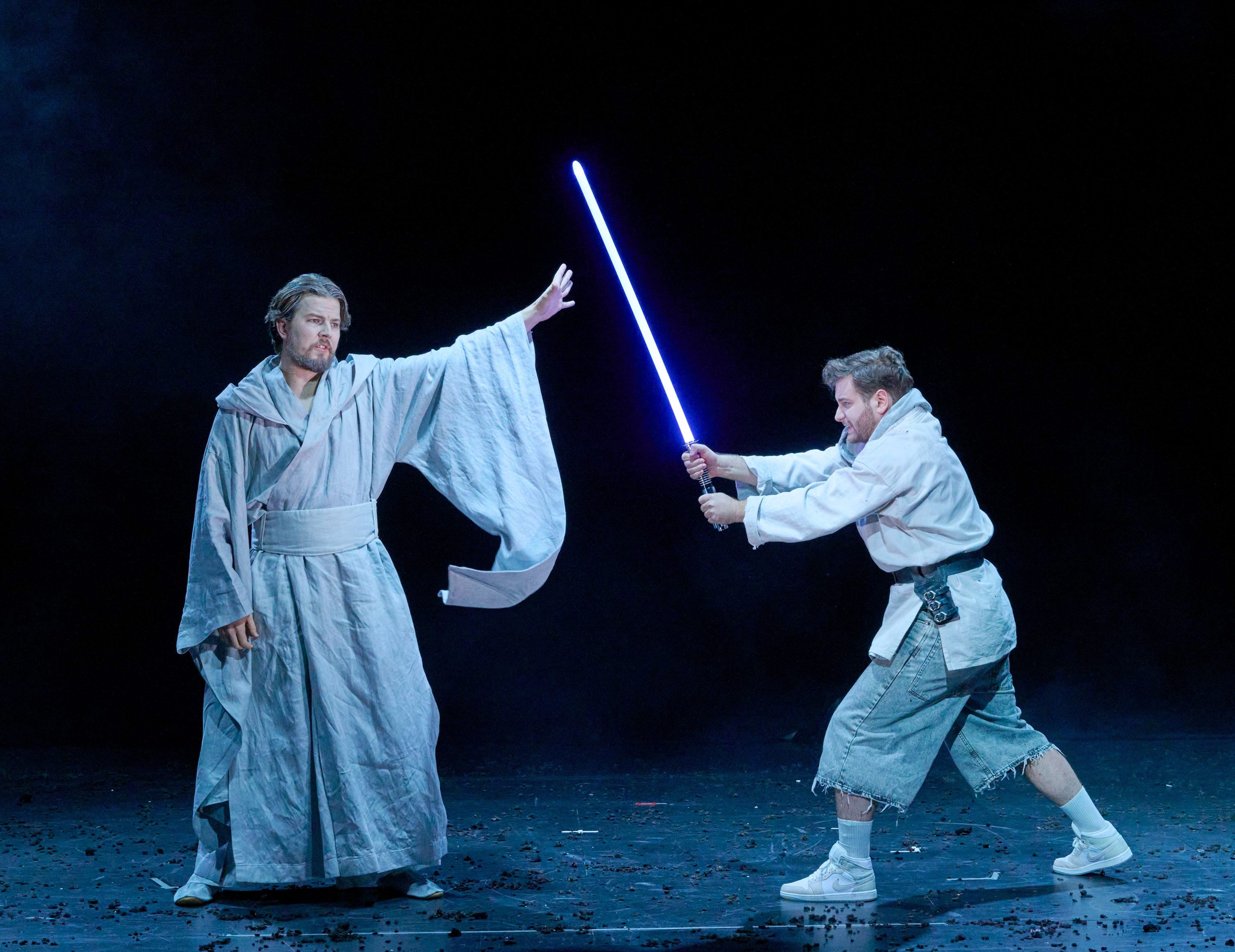 Two actors perform on stage. One, wearing a flowing gray robe, extends a hand as if using a power, while the other, in casual attire with a lightsaber, appears ready for combat. The background is dark, enhancing the dramatic scene.